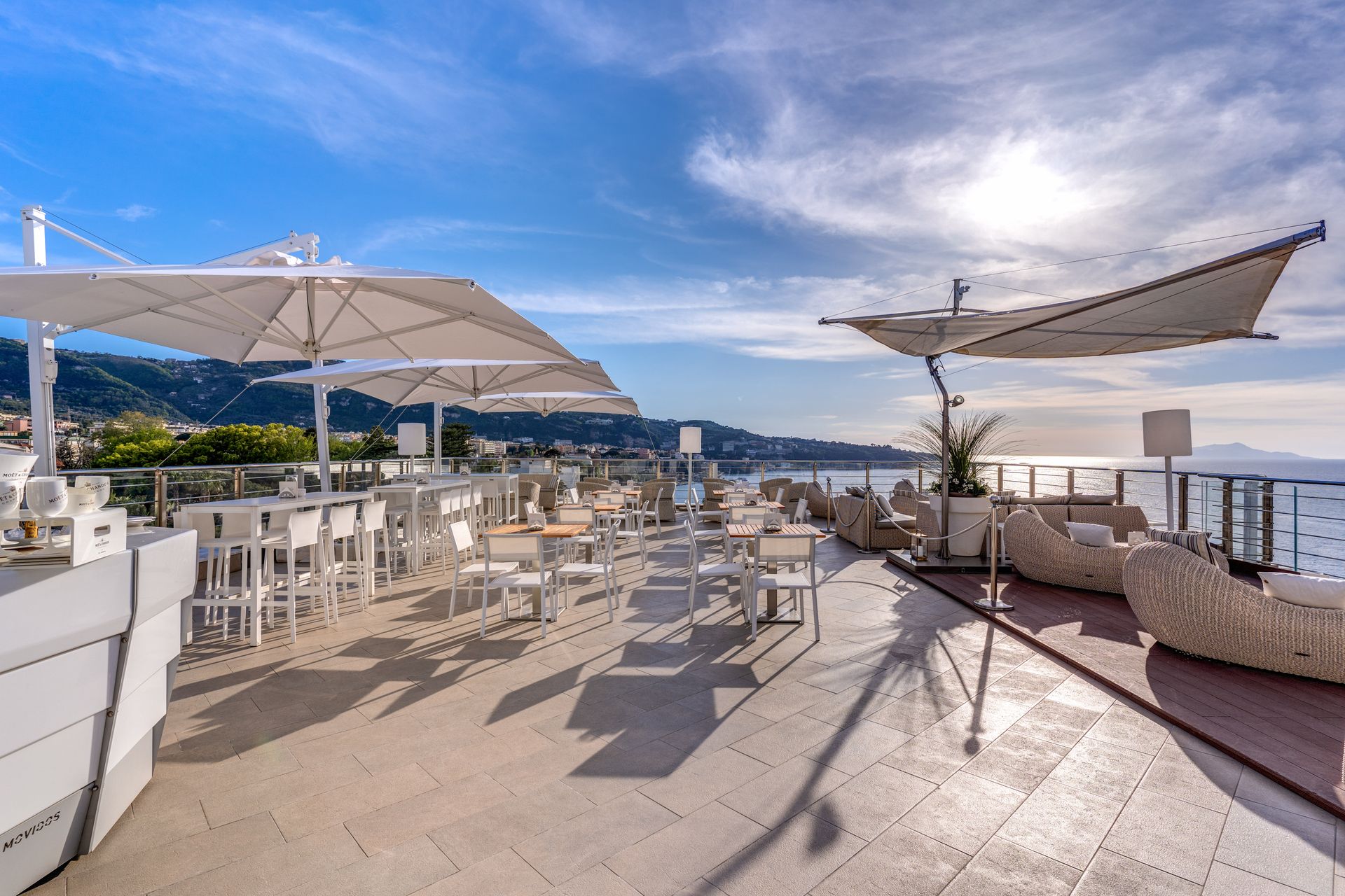 Una terrazza panoramica all'aperto con tavoli, ombrelloni bianchi e lettini prendisole che si affaccia sul mare sotto un cielo azzurro e soleggiato.