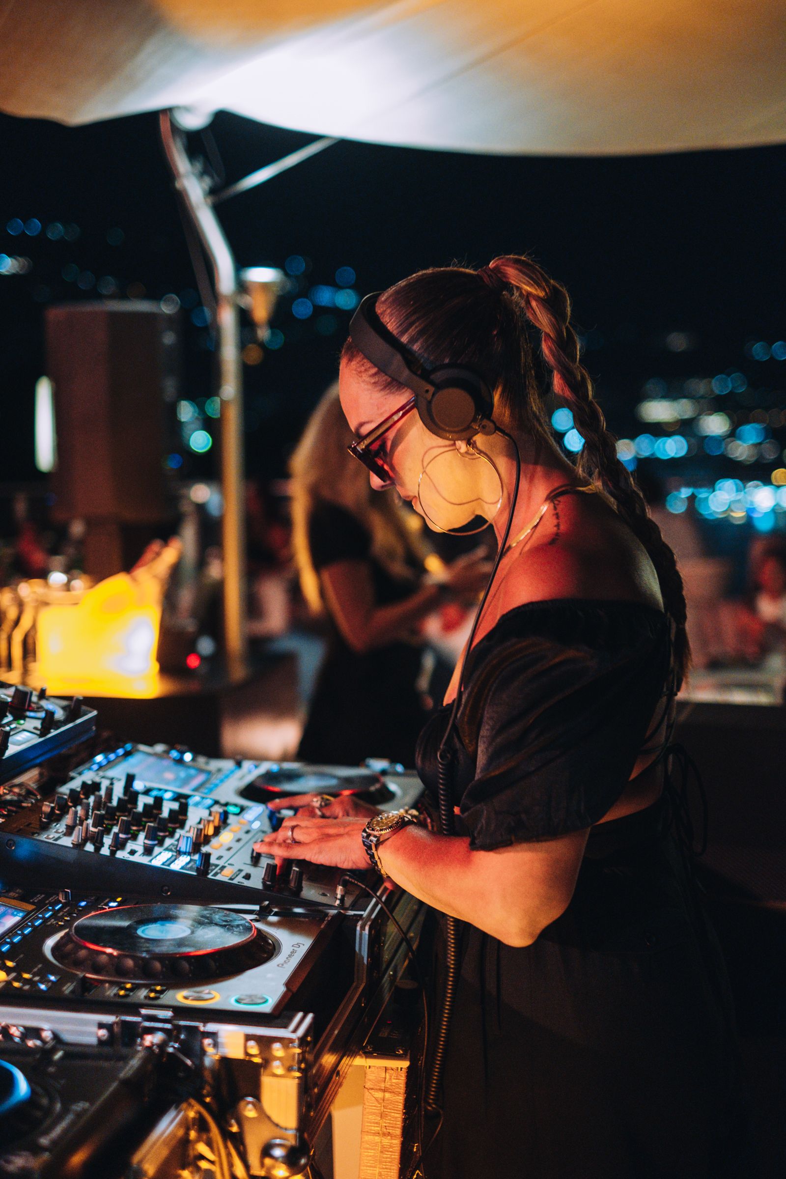 Una persona con i capelli intrecciati, che indossa cuffie e occhiali da sole, mentre fa la DJ a un evento notturno all'aperto.