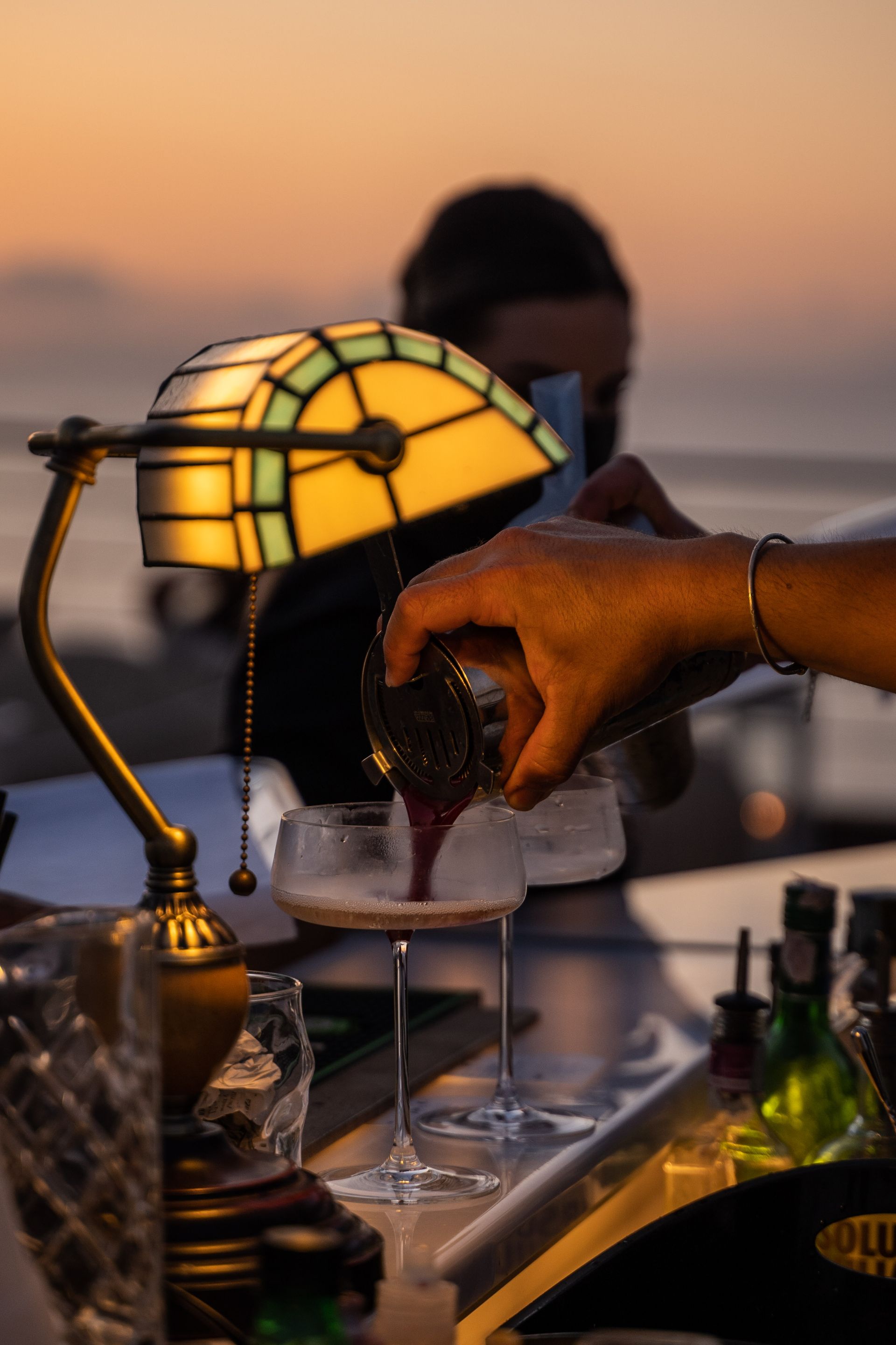 A bartender pours a dark cocktail into a coupe glass at a bar at sunset, illuminated by a classic bankers desk lamp.