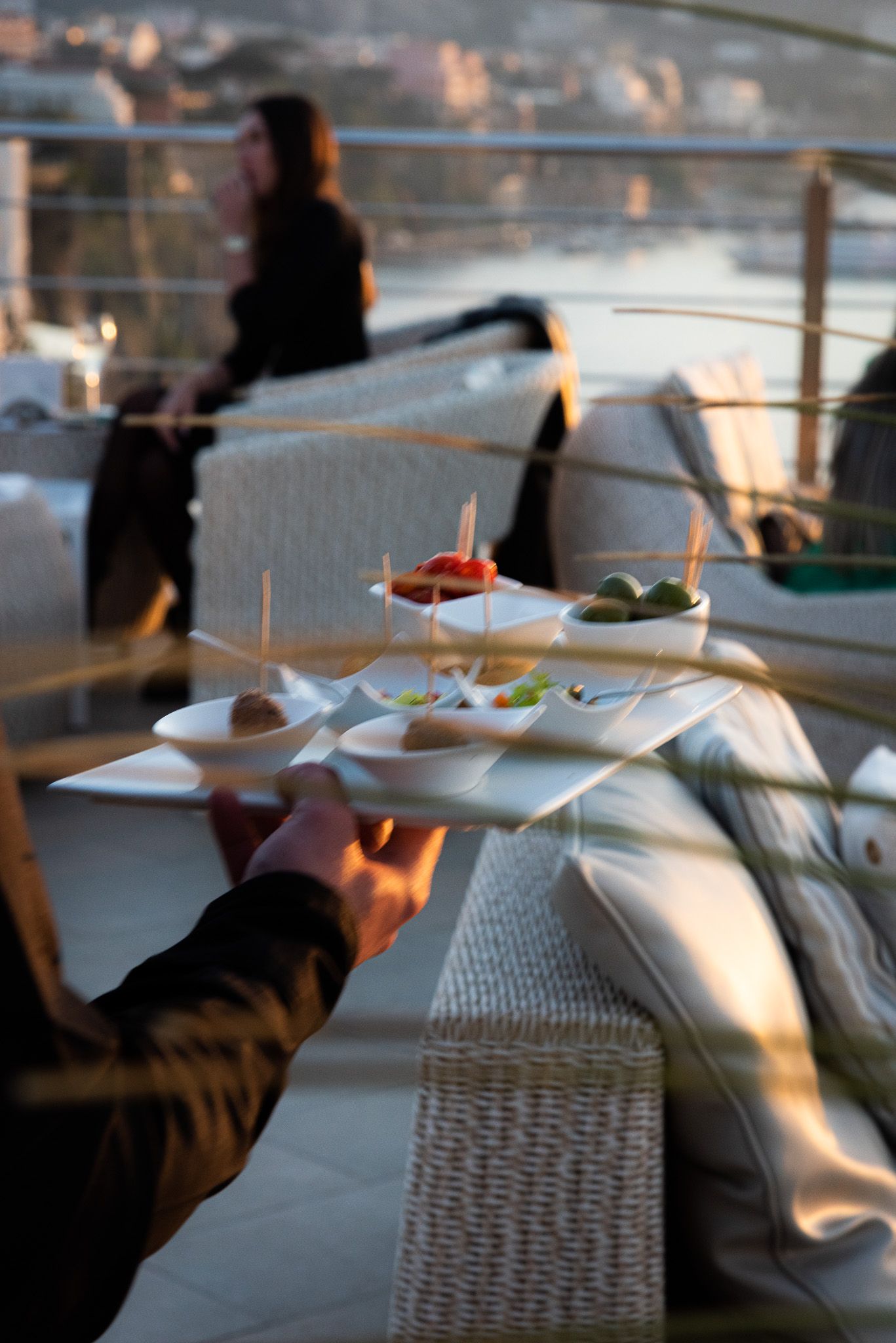 In un locale panoramico sul tetto, al tramonto, un cameriere tiene in mano un vassoio con delle piccole ciotole di antipasti, mentre nelle vicinanze è seduta una persona sfocata.