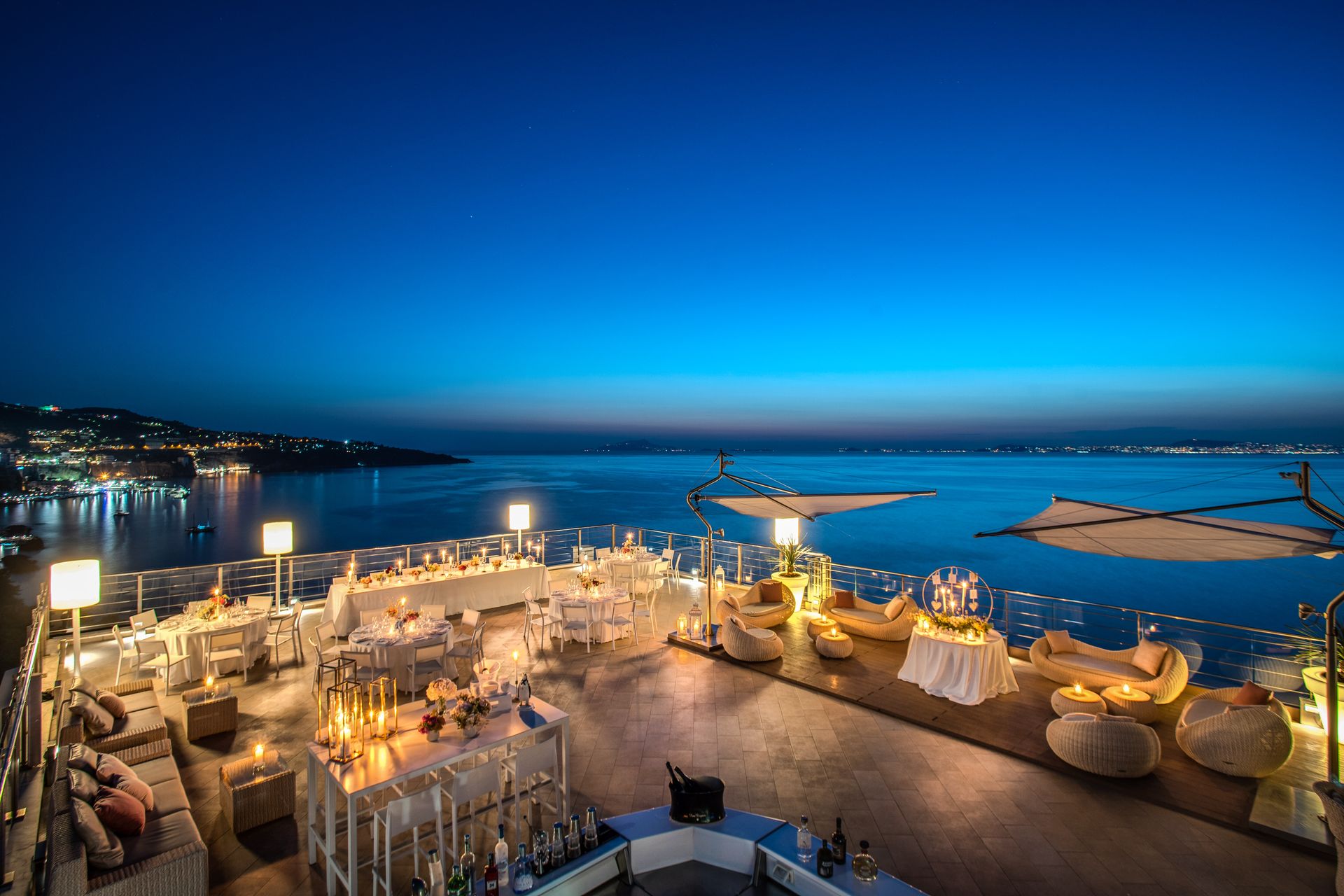 Una vista del tramonto dalla terrazza all'aperto di un ristorante affacciato sul mare, con tavoli da pranzo e comode poltrone.