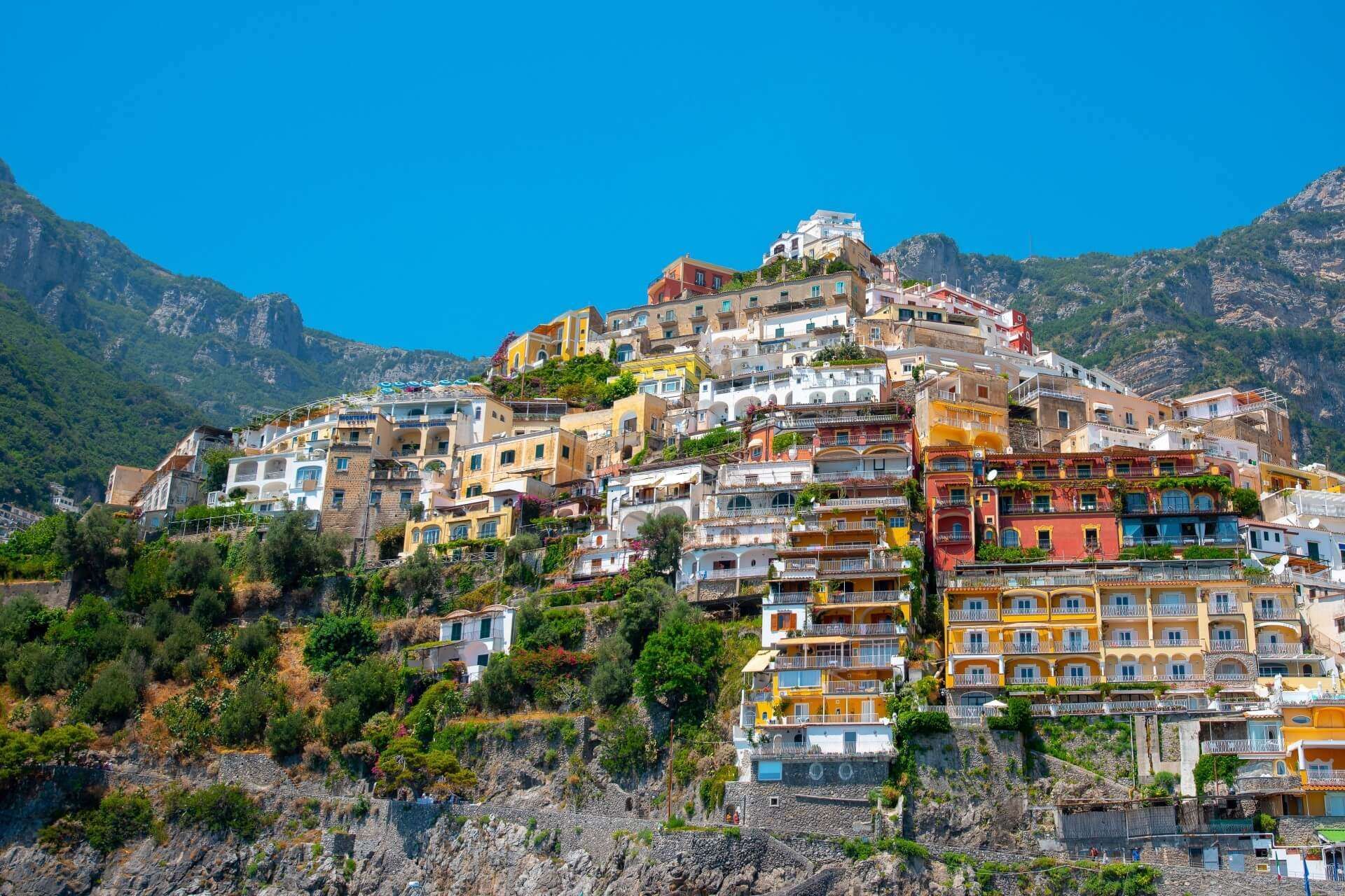Colorful villas cascade down a steep, sunny hillside overlooking the coast in Positano, Italy, under a clear blue sky.