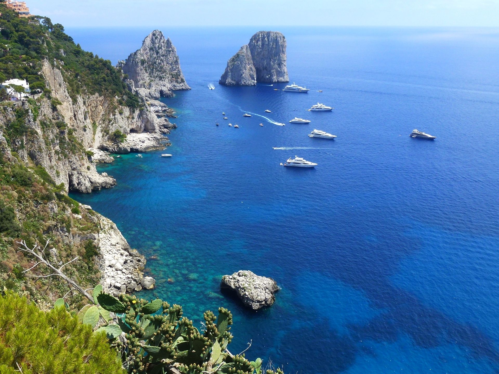 High cliffs and rock formations rise from deep blue Mediterranean water, populated by several small white boats near Capri.