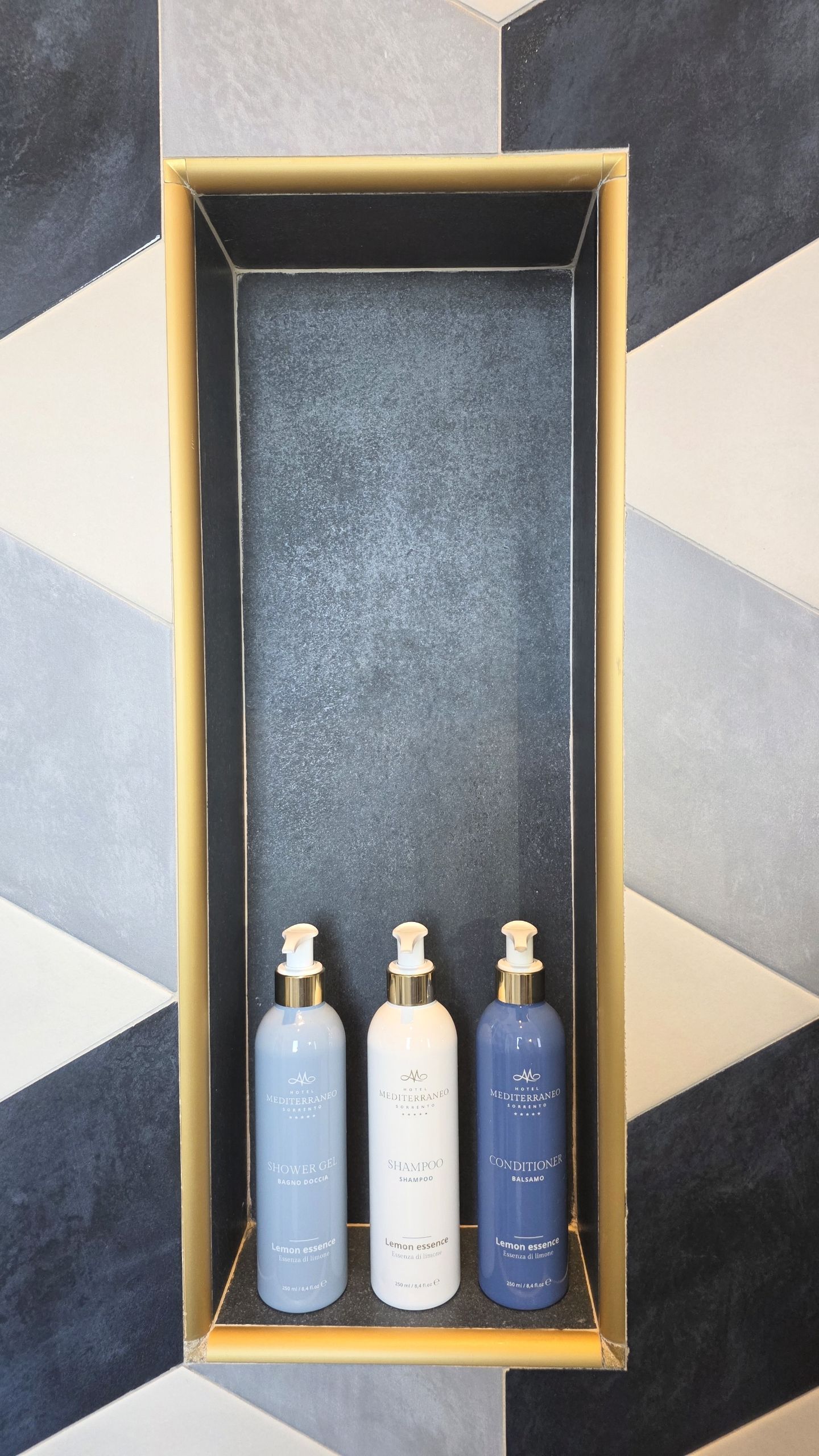 Tre flaconi di sapone con etichette decorative sono collocati in una nicchia rettangolare della doccia con cornice dorata, addossati a una parete rivestita di piastrelle geometriche.