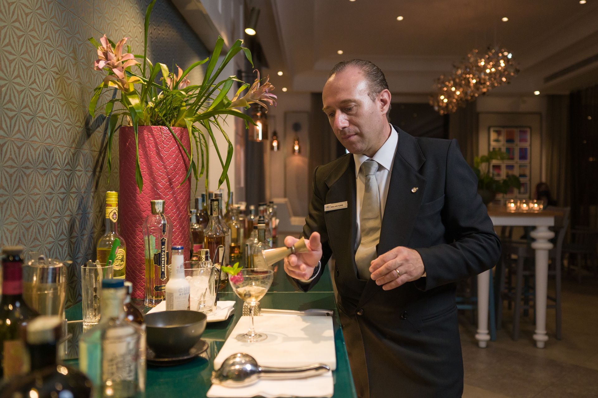 Un barista professionista in abito scuro prepara un cocktail al bancone di un bar, con un vaso alto e diverse bottiglie.