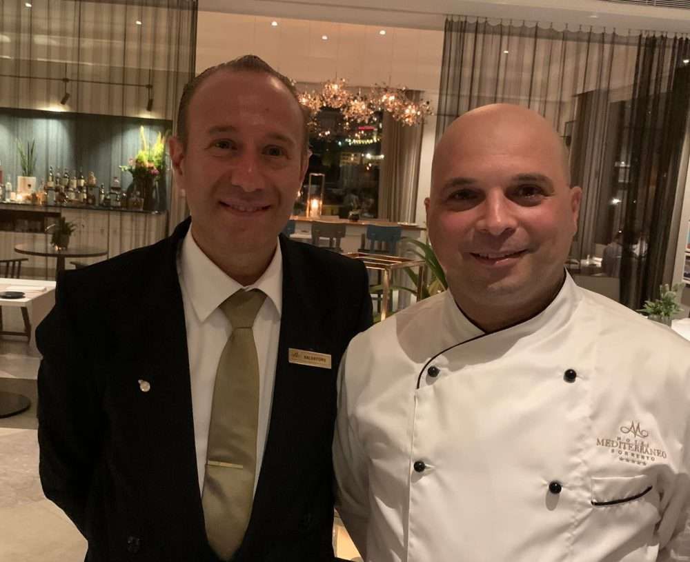 Two men in professional attire—one in a suit and tie, the other in a chef's uniform—stand together in a restaurant.