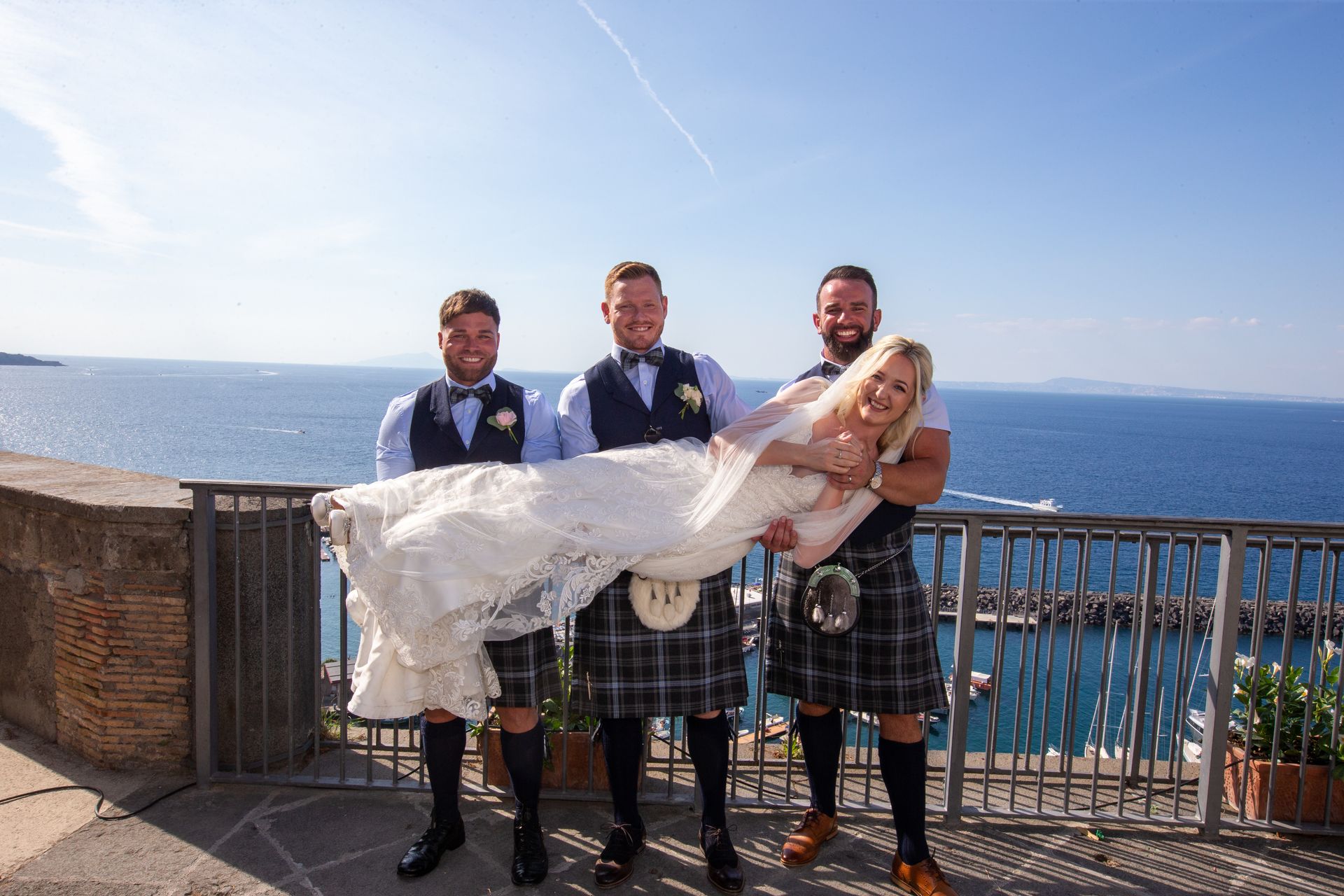 Tre uomini in abiti tradizionali scozzesi sorreggono orizzontalmente una sposa in abito da sposa di pizzo bianco, con lo sfondo dell'oceano.