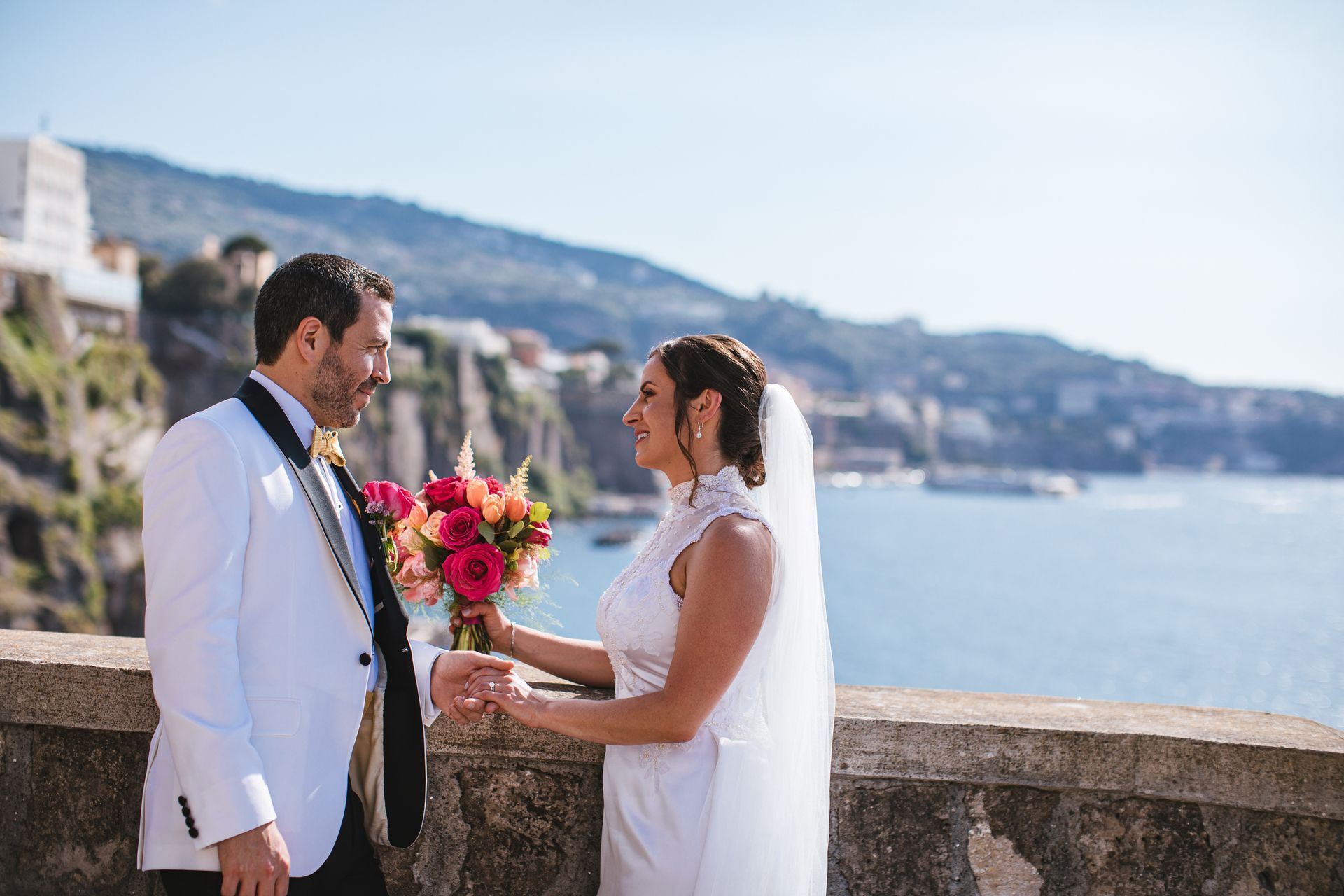 A bride and groom in wedding attire hold a colorful bouquet while facing each other on a balcony overlooking the sea.