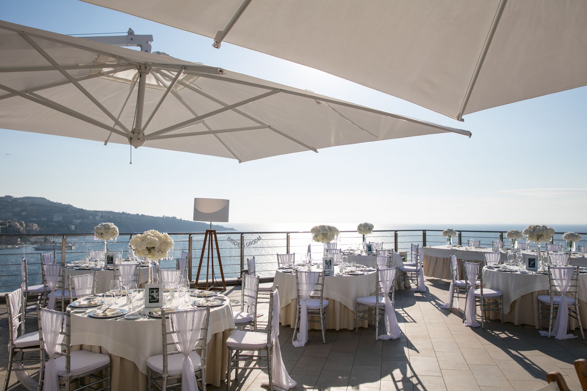 Ricevimento di nozze all'aperto su una terrazza con vista sull'oceano, con tavoli rotondi, sedie bianche e grandi ombrelloni.