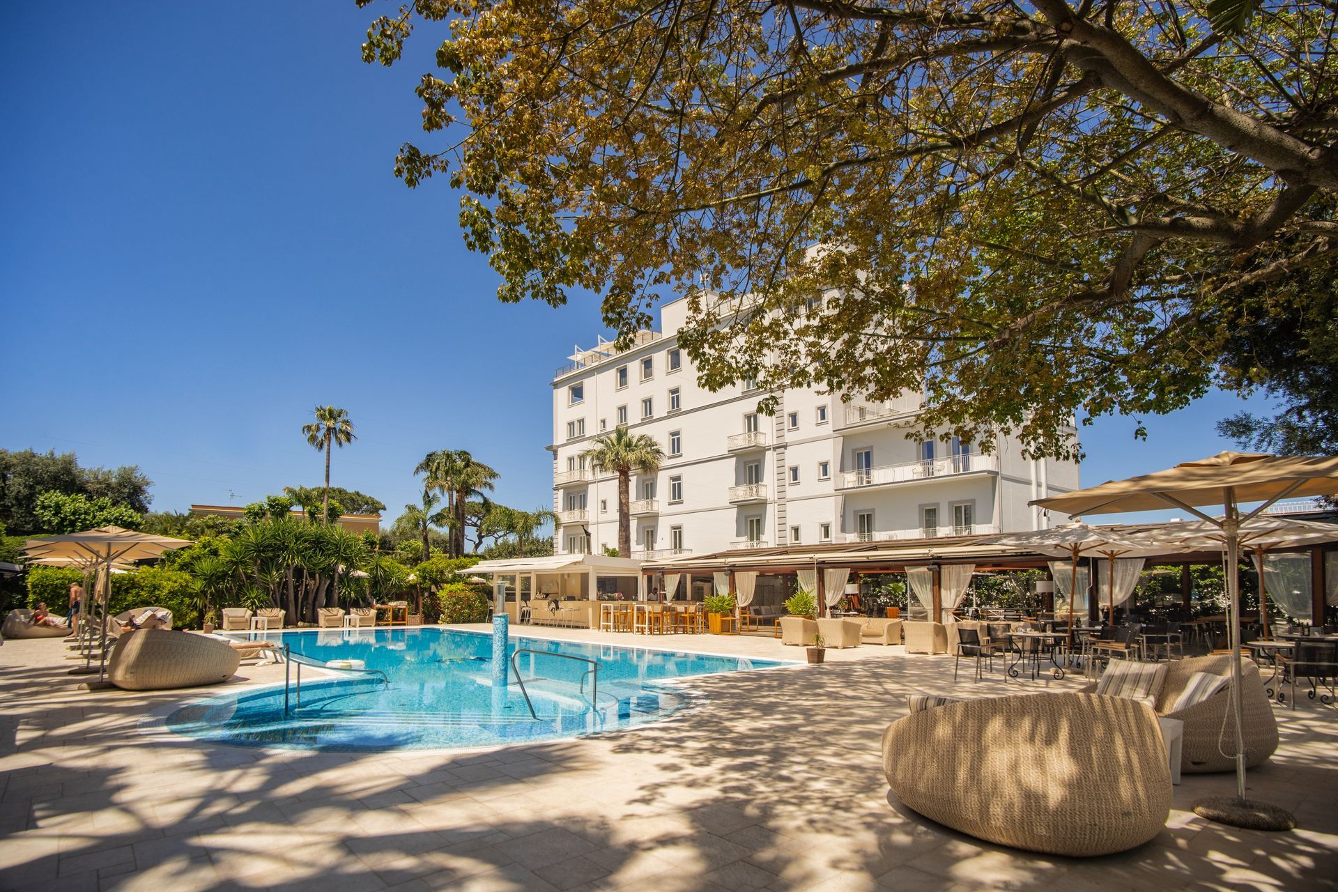A sunny outdoor hotel pool area with a white multi-story building, palm trees, lounge chairs, and shaded dining tables.