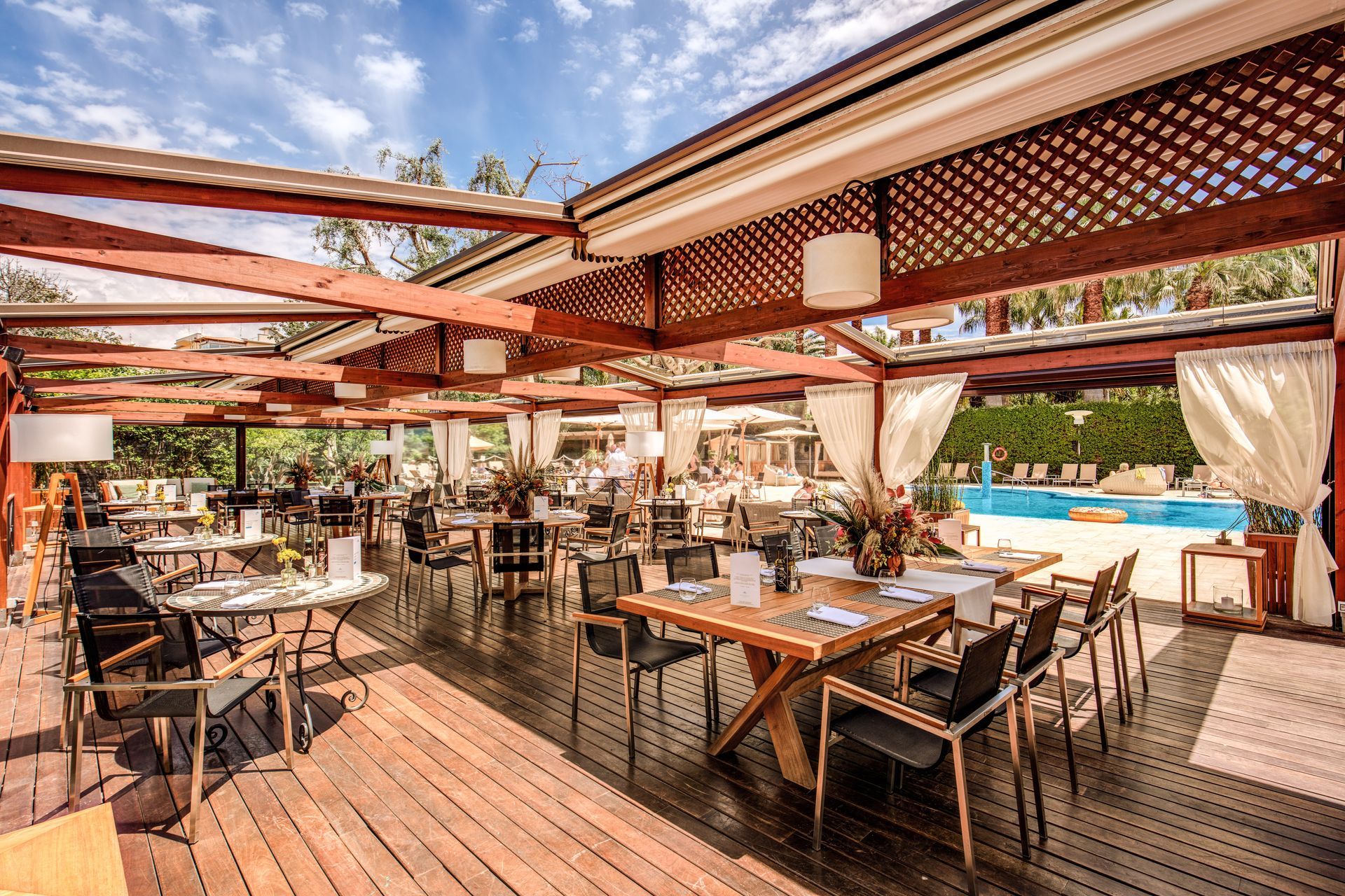 Terrazza esterna del ristorante con tavoli in legno, sedie intrecciate, pergola rossa e vista sulla piscina.