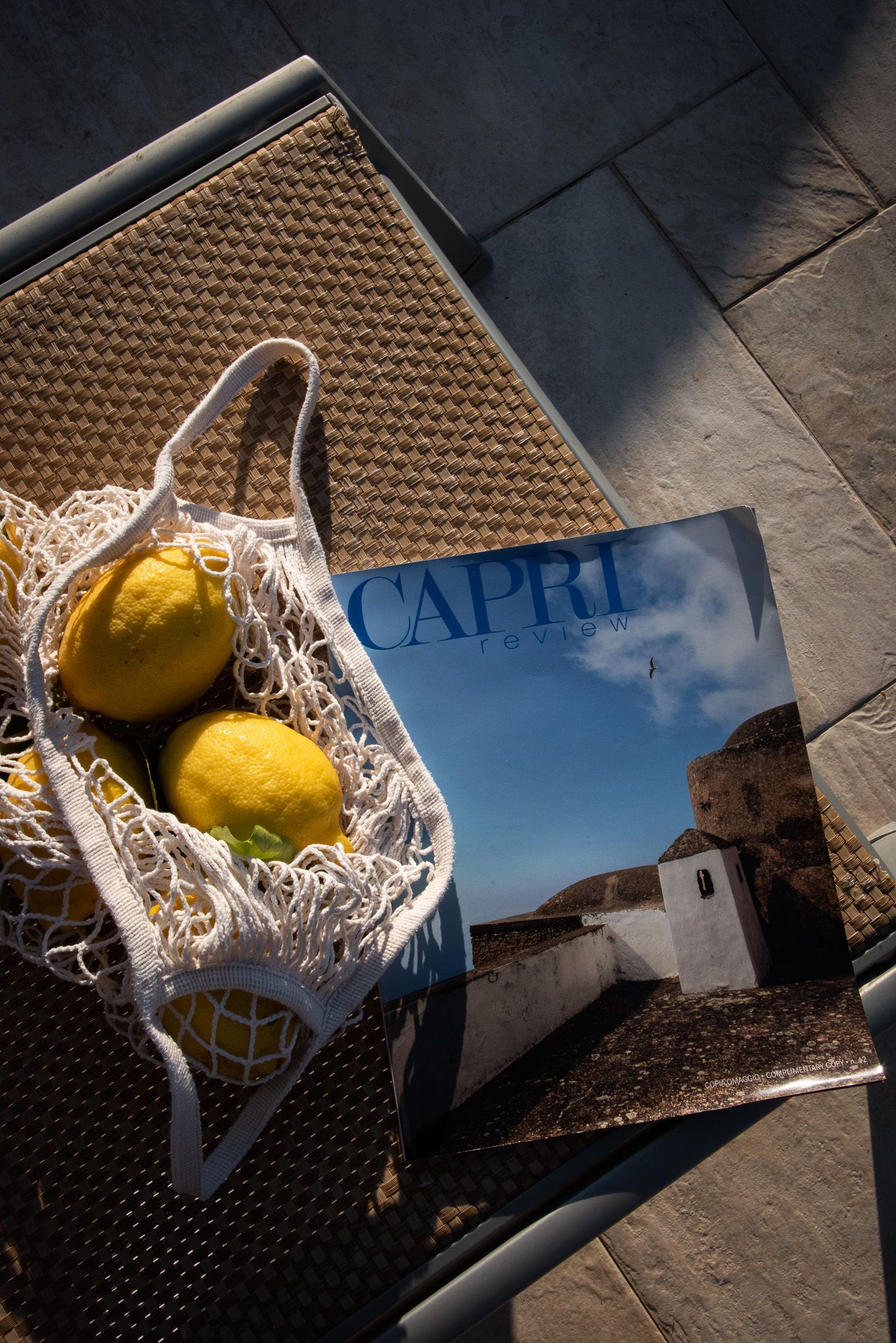 Una borsa all'uncinetto piena di limoni giallo brillante è appoggiata su una superficie ruvida accanto a un libro di viaggi intitolato