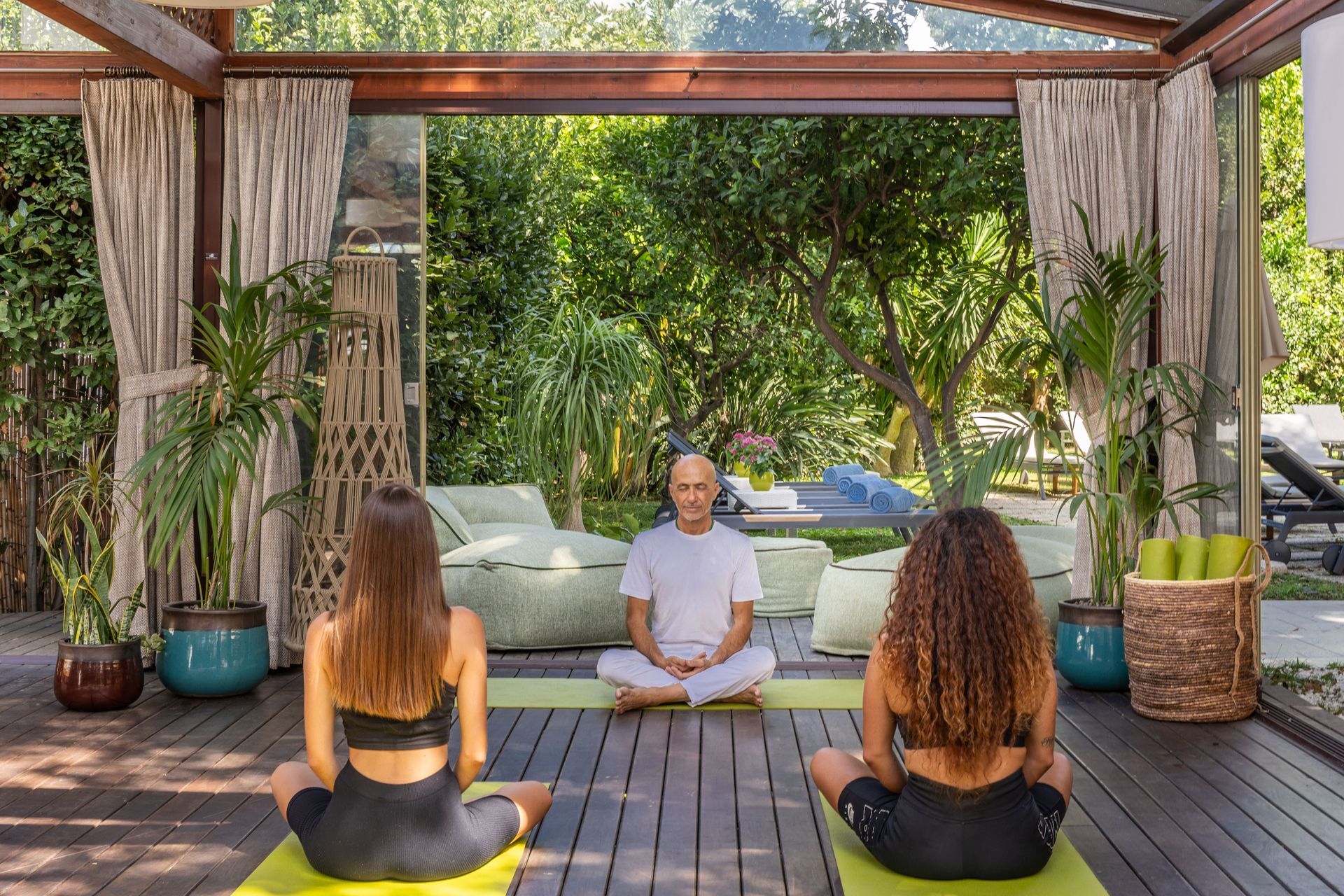 Three people sit in meditation poses on a wooden deck in a sunlit, open-air pavilion surrounded by lush greenery.