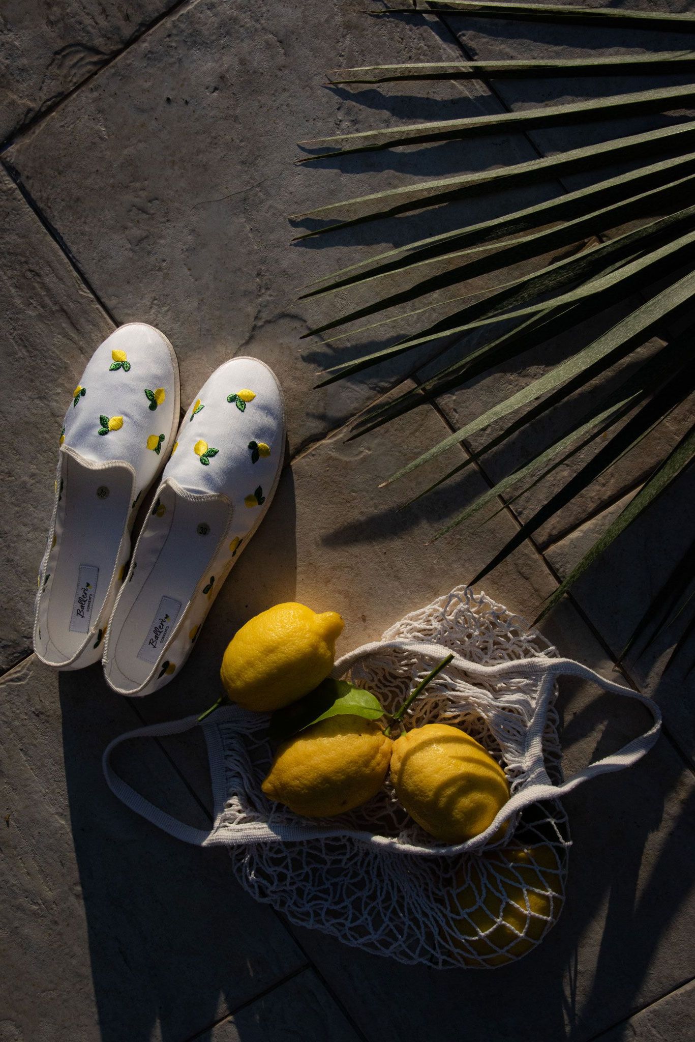 A pair of white lemon-print espadrilles next to a mesh bag containing three fresh lemons on a stone patio with palm leaves.