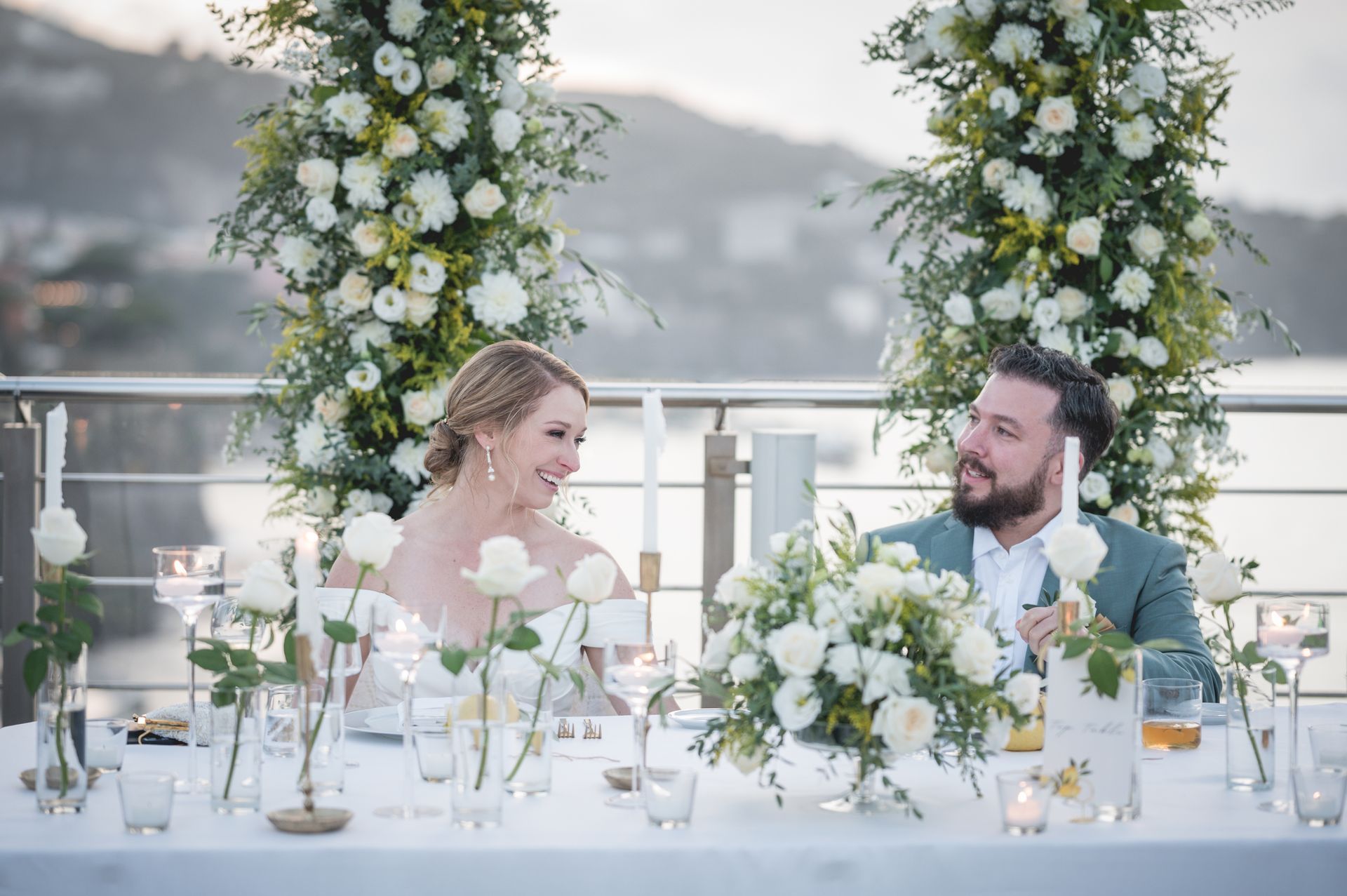 A couple shares a happy gaze at their outdoor wedding reception, seated at a table adorned with white flowers and candles.