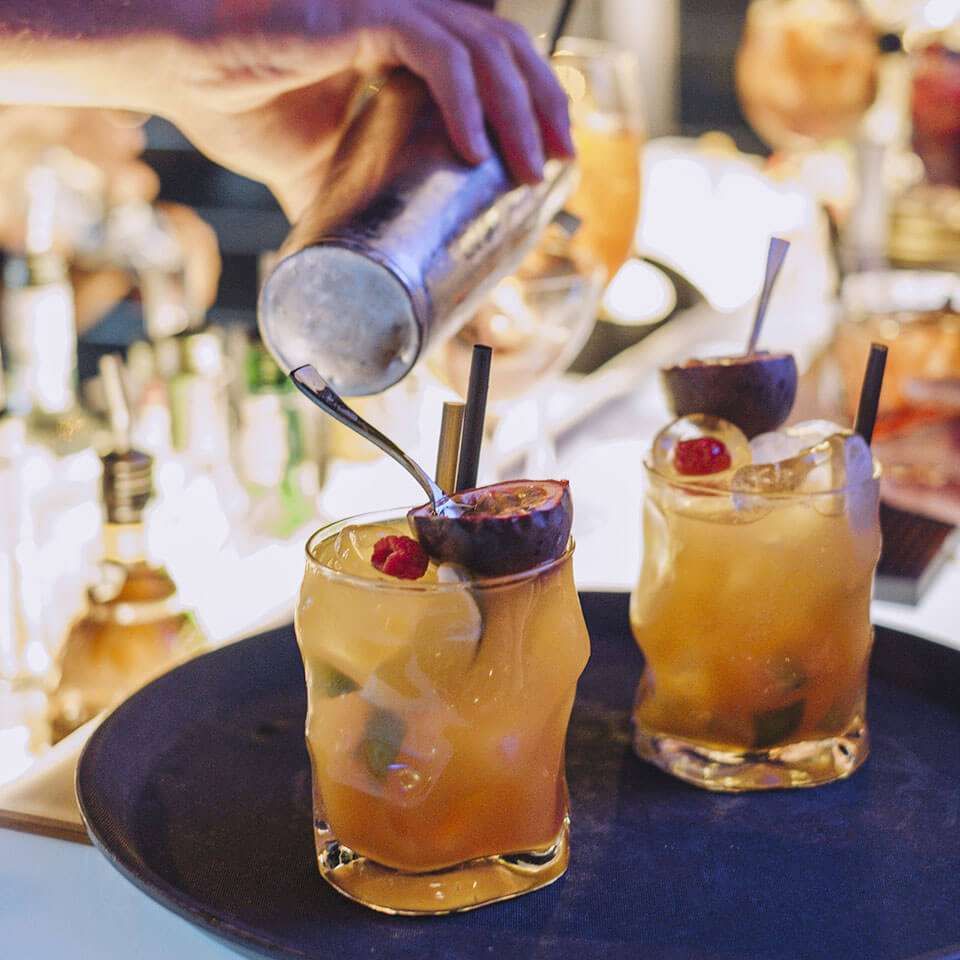 A bartender pours a drink into glasses garnished with passion fruit and berries on a tray at a bar.