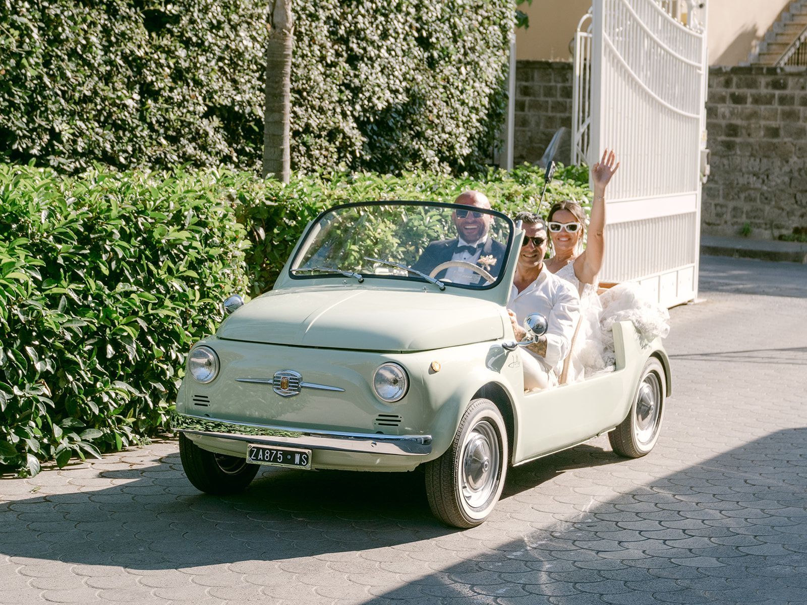 A couple in formal wedding attire kisses beside a white convertible in front of a white building with arched doorways.