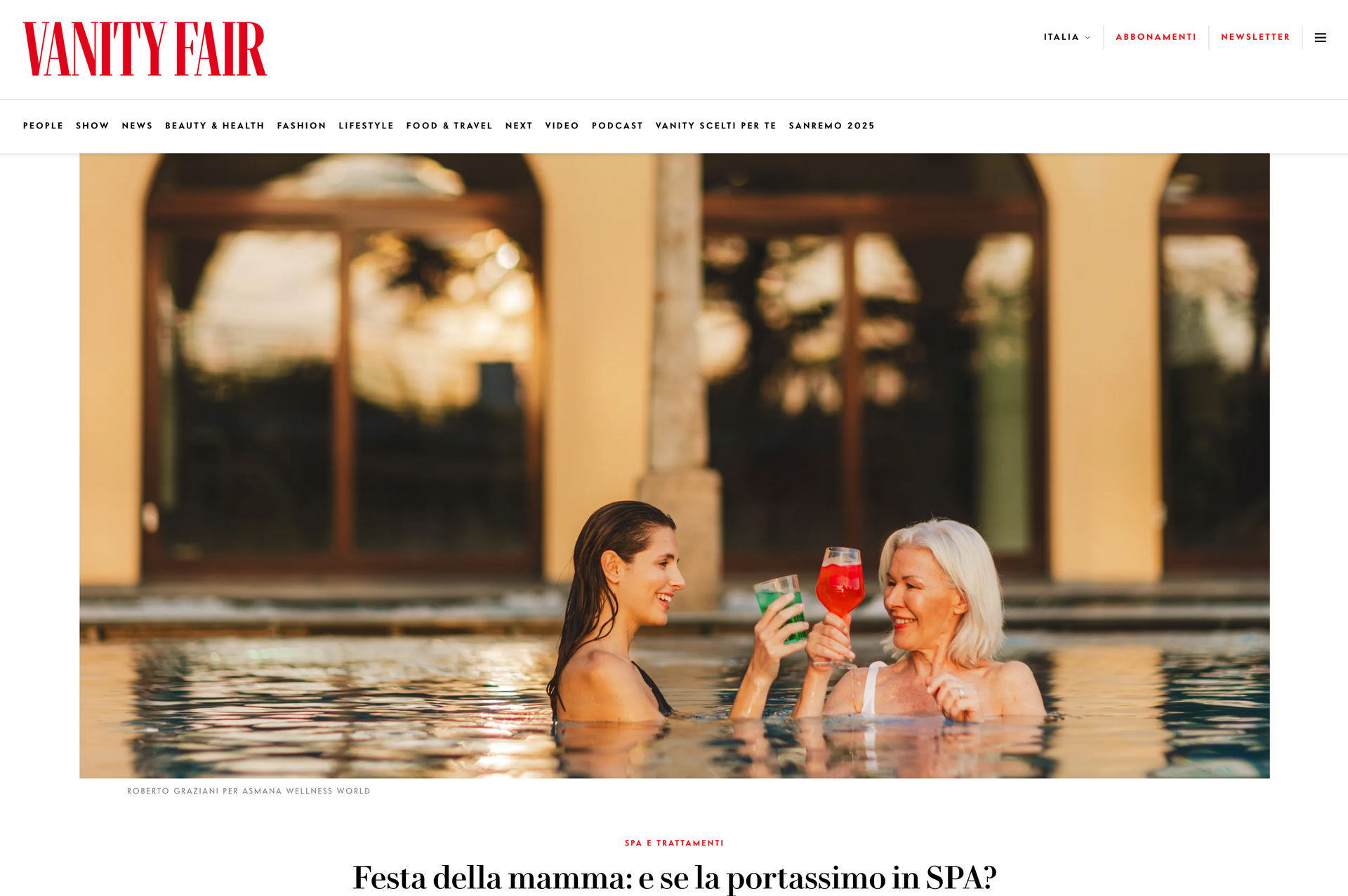 Two women clink glasses in a swimming pool at sunset in front of a building with arched windows.