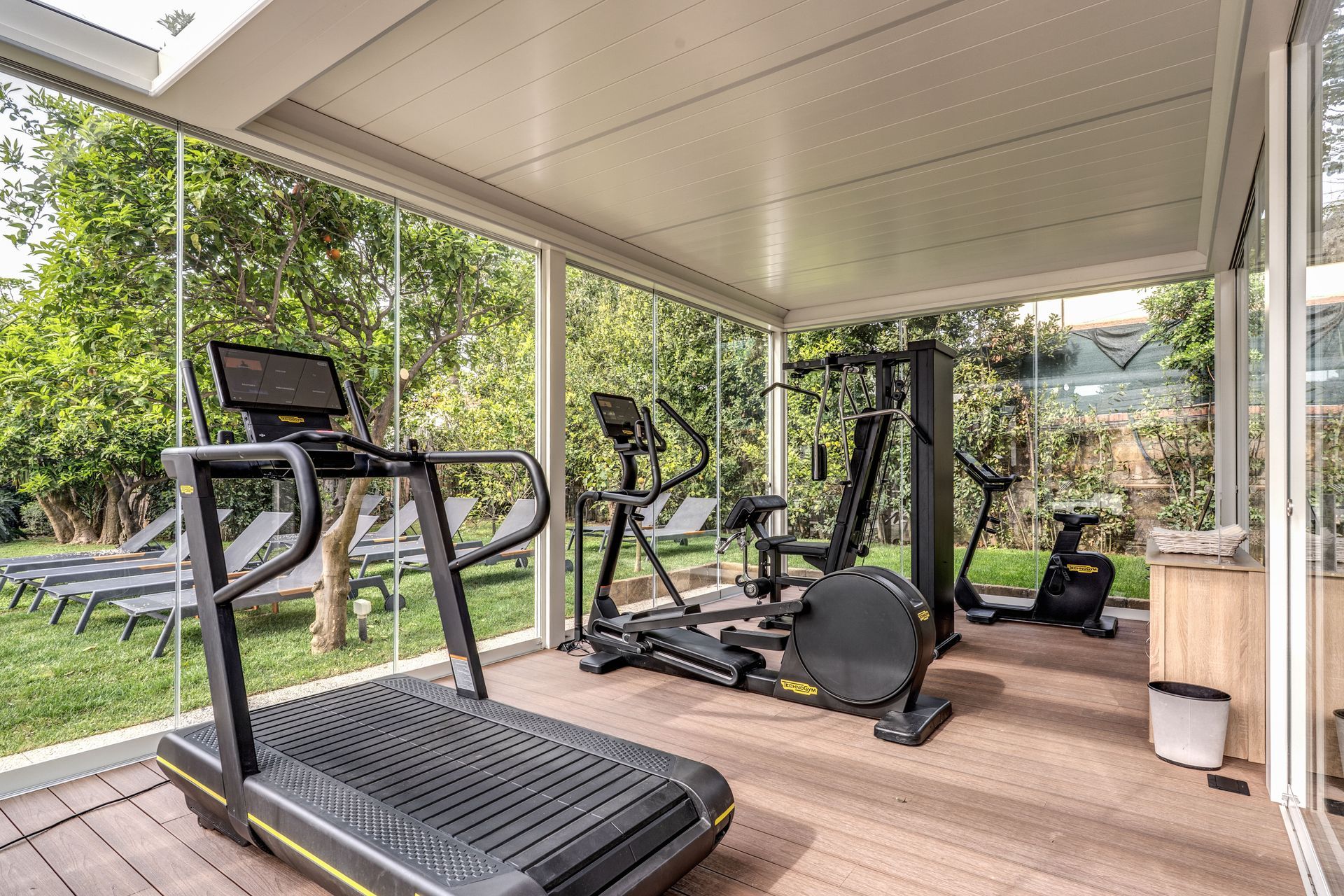 A modern, glass-walled gym featuring a treadmill, elliptical, multi-gym station, and stationary bike on a wooden deck.