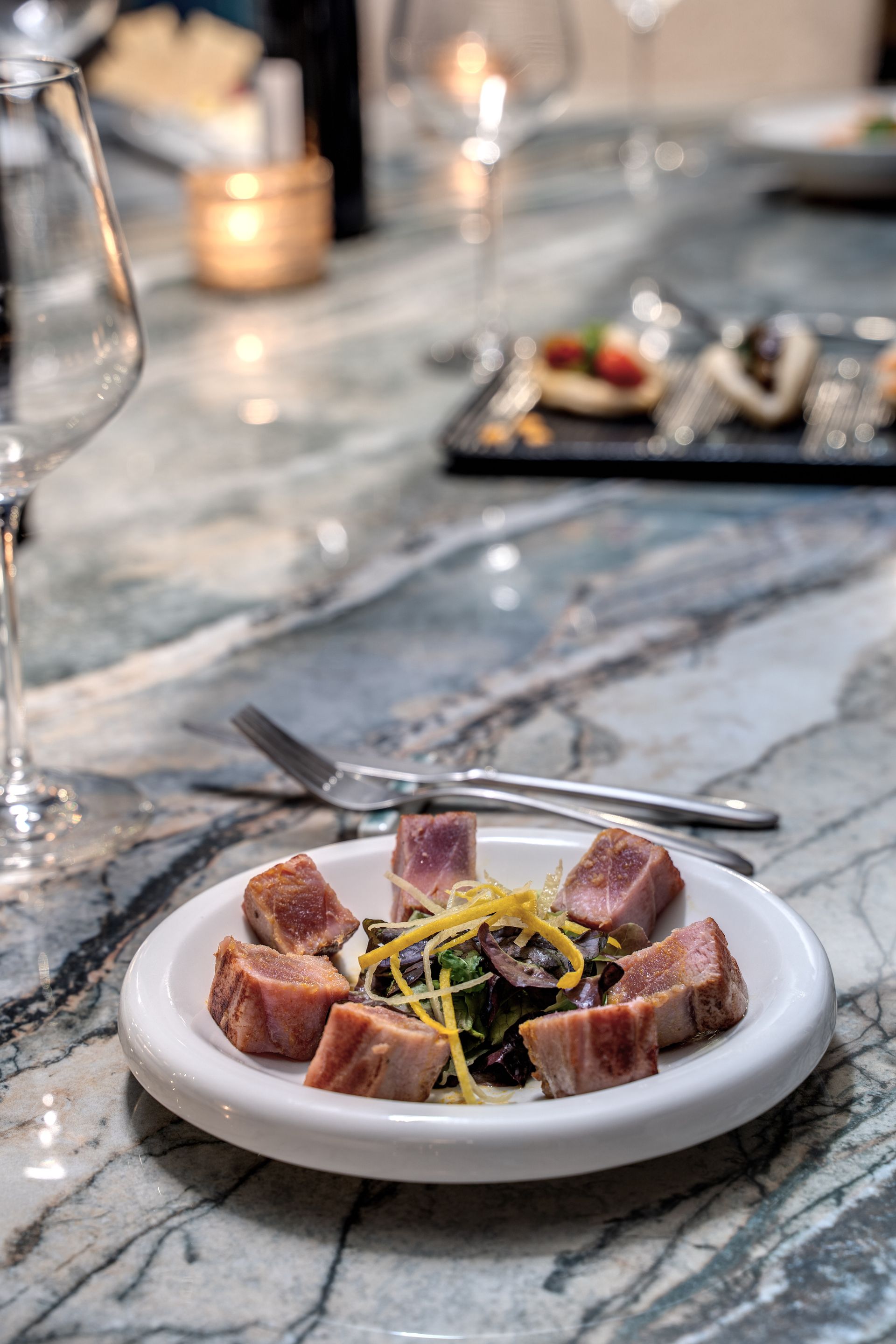 A plate of seared tuna chunks with garnishes on a marbled table in a dimly lit restaurant.