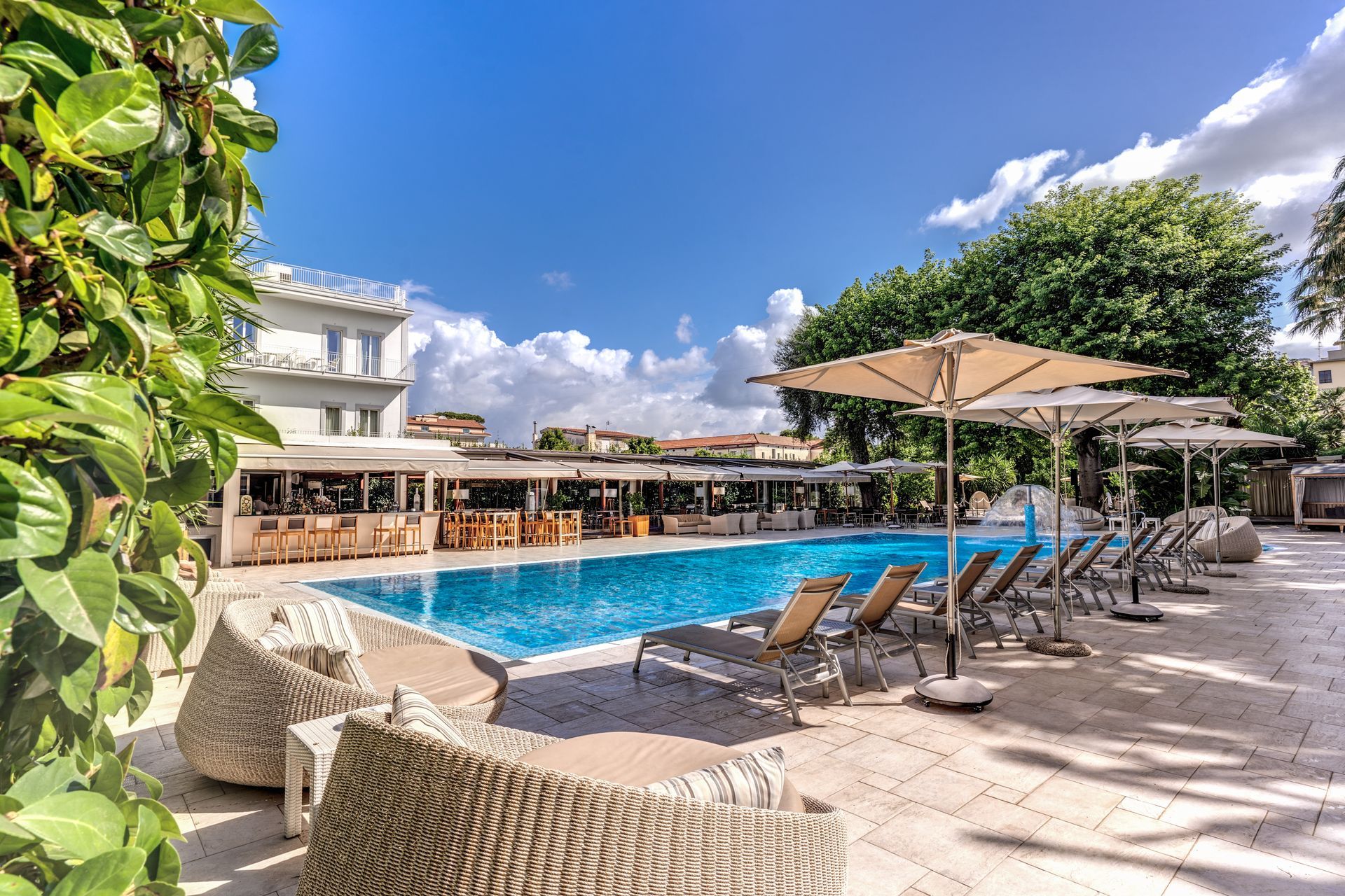 Il patio della piscina di un hotel, illuminato dal sole, è caratterizzato da acque azzurre, file di lettini con ombrelloni e piastrelle di pietra bianca, il tutto sotto un cielo azzurro.