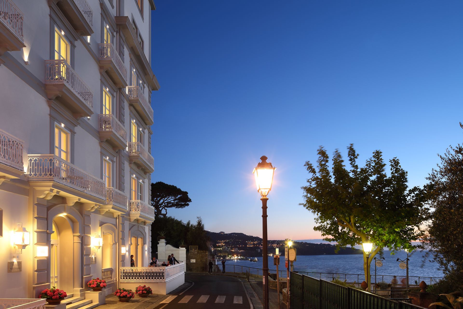 Al crepuscolo, la facciata illuminata dell'hotel si affaccia sul mare, con i lampioni che brillano lungo una passeggiata a ridosso dell'acqua.