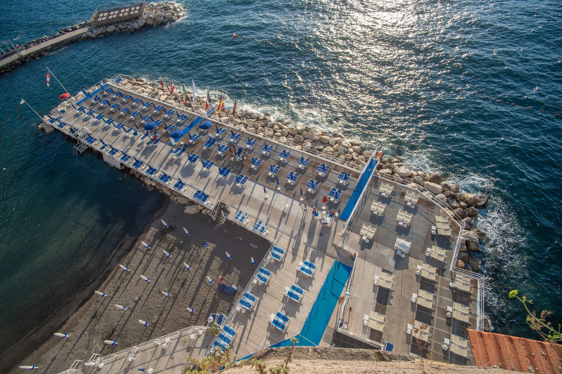 An elevated view of a coastal beach club with rows of blue and white sun loungers on a stone pier next to the dark sea.