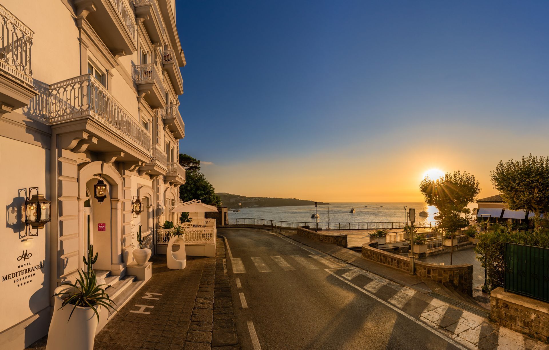 A golden sunset illuminates a coastal road beside a white multi-story hotel with iron balconies overlooking the sea.