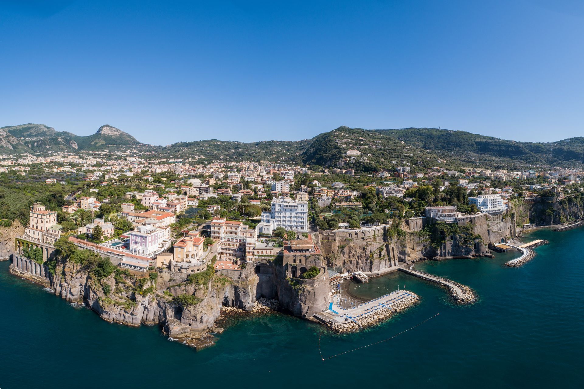 An aerial view of the town of Sorrento, Italy, with white buildings perched on rocky cliffs above the blue Tyrrhenian Sea.