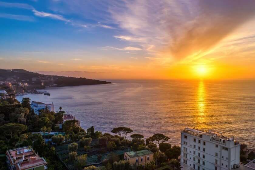 A vibrant sunset over a coastal town, with golden light reflecting on the calm ocean and silhouettes of trees and buildings.