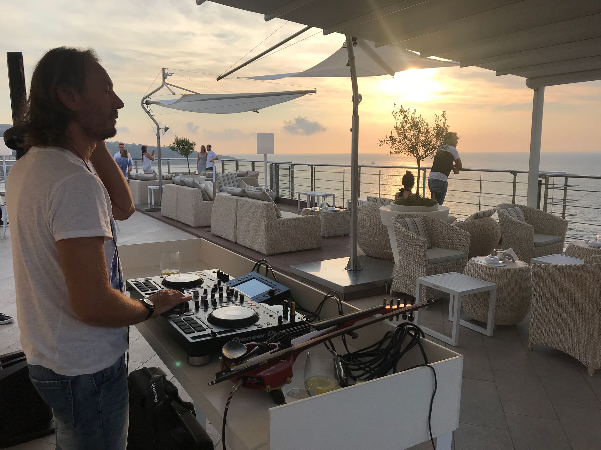 Un DJ mixa musica su una terrazza con vista sull'oceano al tramonto, mentre le persone sono sedute su comodi lettini nelle vicinanze.
