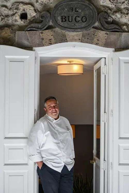 A chef in a white uniform stands smiling in the doorway of a restaurant named Al Buco, framed by white wooden doors.