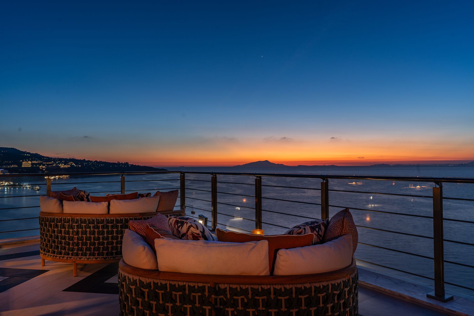 Rooftop lounge chairs facing a sunset over the ocean, with city lights along the coast.