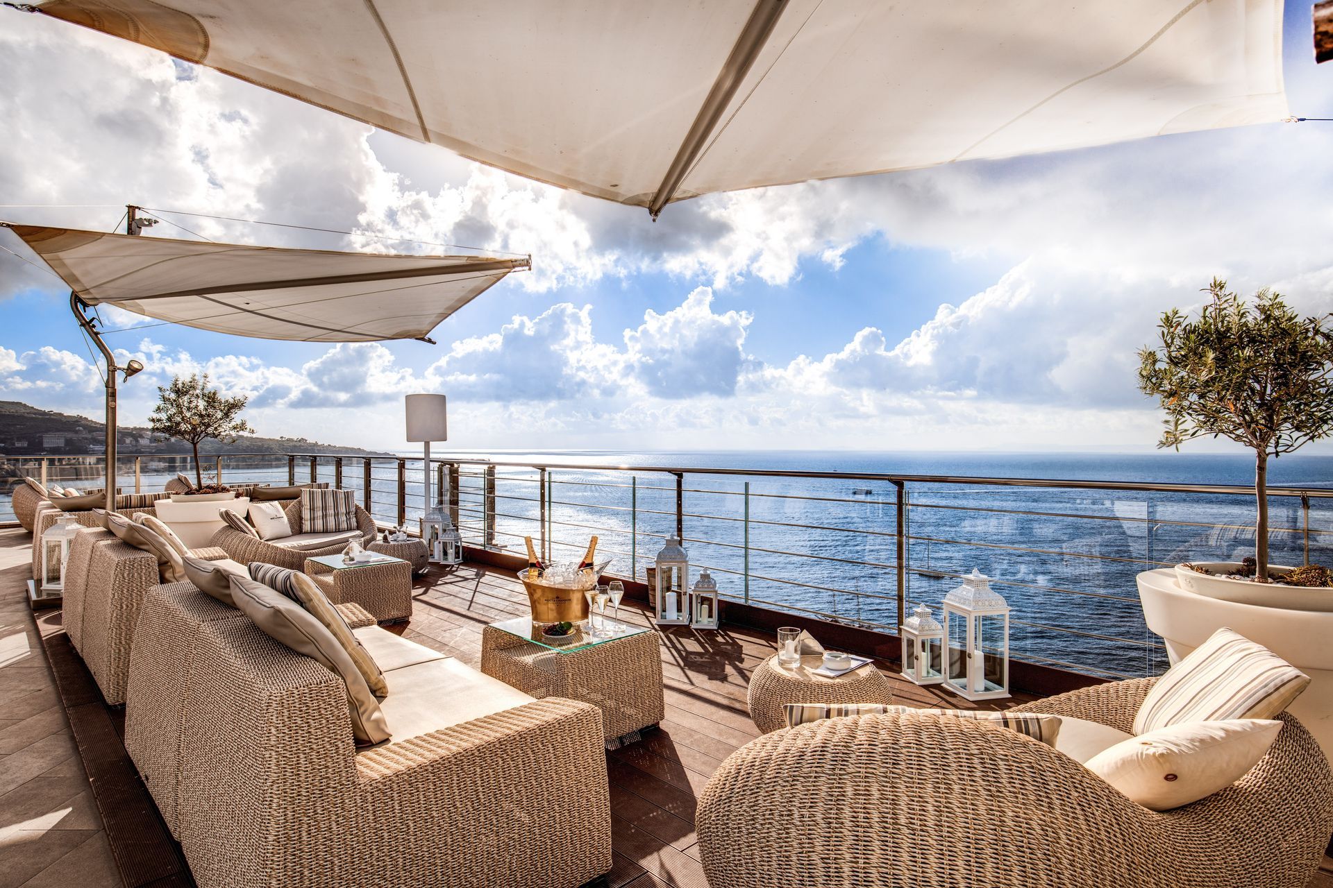 Mobili da salotto in vimini su una terrazza con vista sull'oceano blu, sotto un cielo parzialmente nuvoloso e all'ombra di un baldacchino.