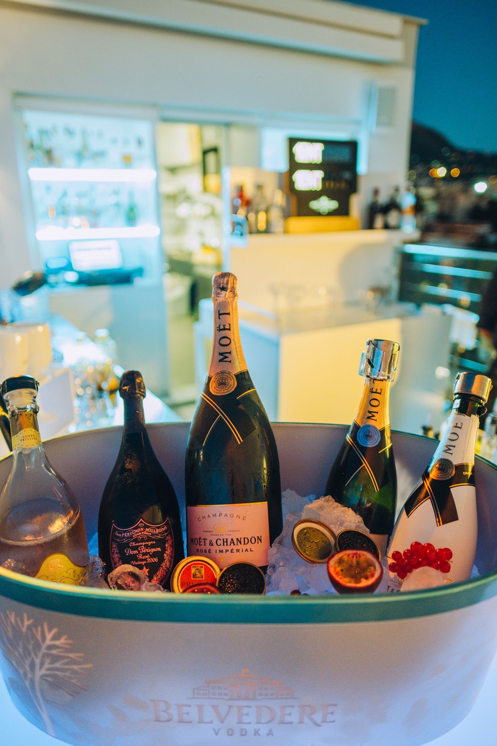 Un grande secchiello per il ghiaccio con il marchio Belvedere, pieno di bottiglie di champagne e frutti della passione, in un bar all'aperto di sera.