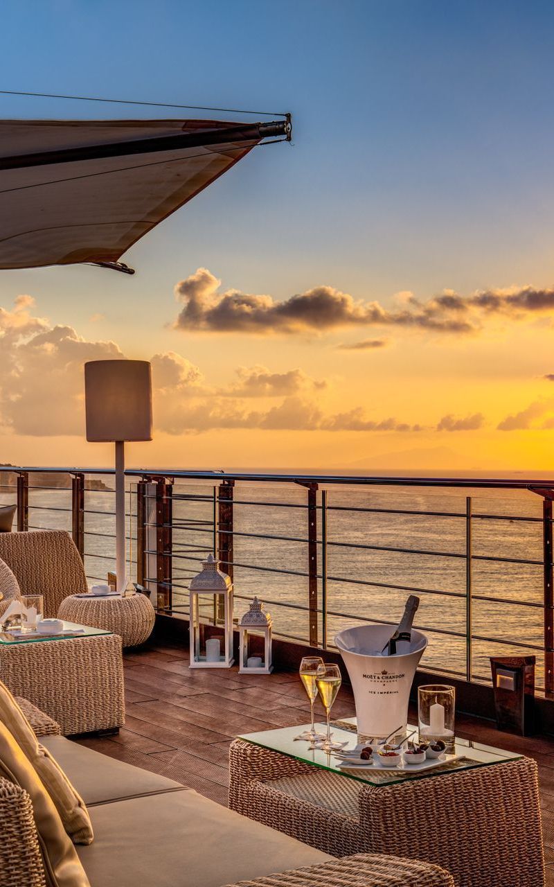 Un tramonto suggestivo sull'oceano, ammirato da una lussuosa terrazza illuminata a lume di candela, con mobili in vimini e un secchiello per lo champagne.