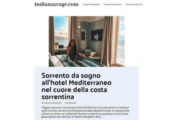A blogger sits on a bed in a hotel room with a view of the sea, featured in an article titled 