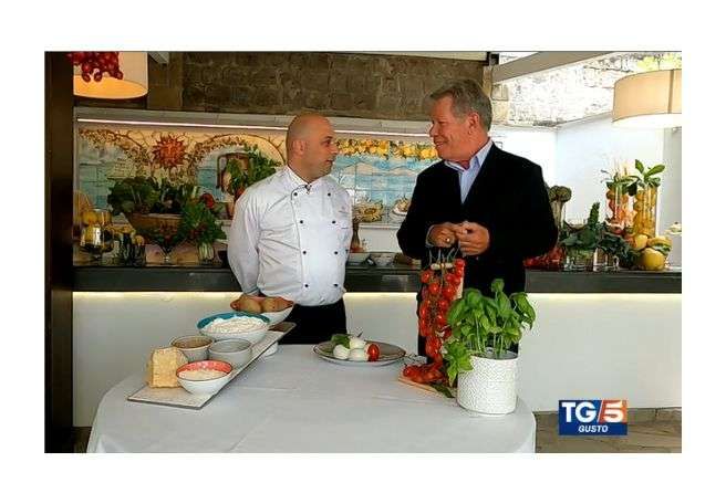 A chef and a man in a suit talk behind a round table with food ingredients, set against a decorated, tiled kitchen wall.