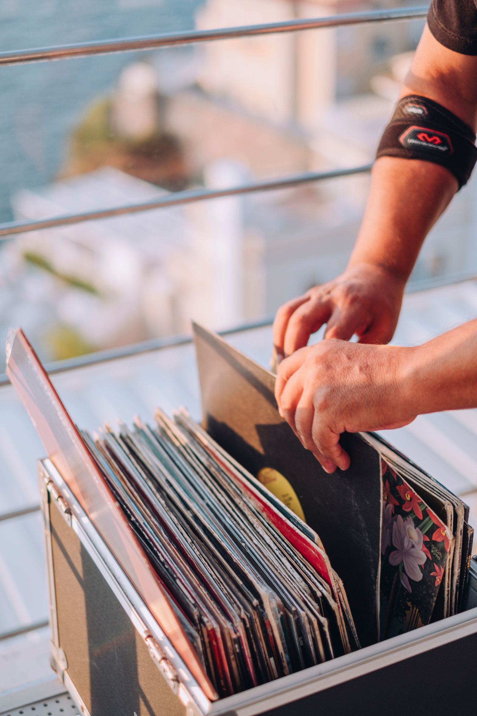 Le mani di una persona sfogliano una collezione di dischi in vinile in una custodia su un balcone affacciato sul mare.