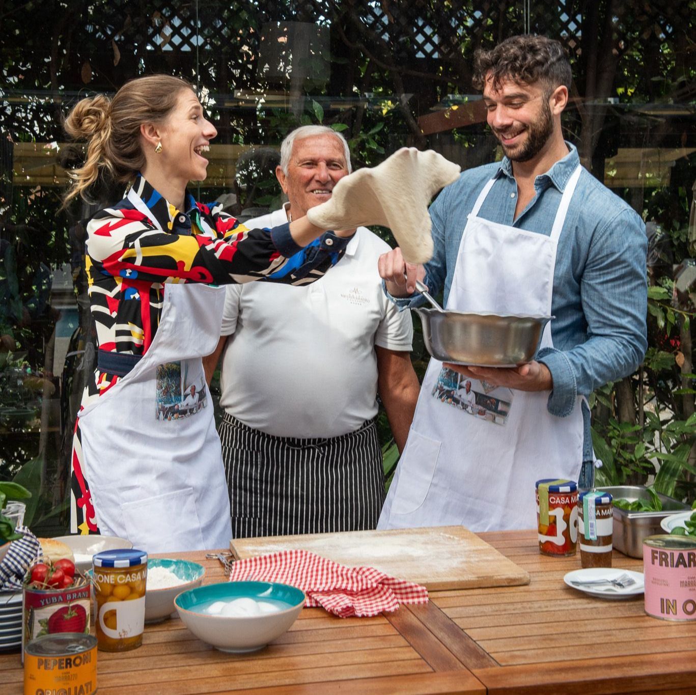Three people in aprons cooking outdoors, smiling while mixing ingredients at a wooden table.