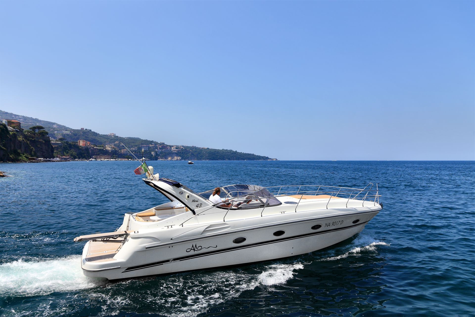A sleek white speedboat cuts through blue sea along a hilly coastline on a sunny day.