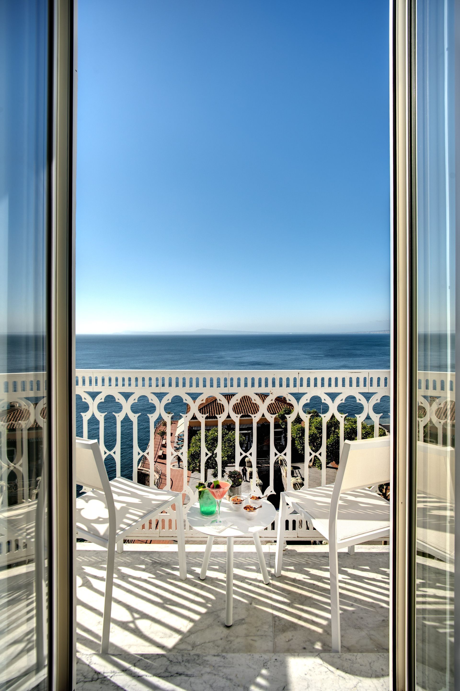 Un balcone bianco affacciato sull'oceano blu, arredato con due sedie e un tavolino, incorniciato da porte scorrevoli in vetro.