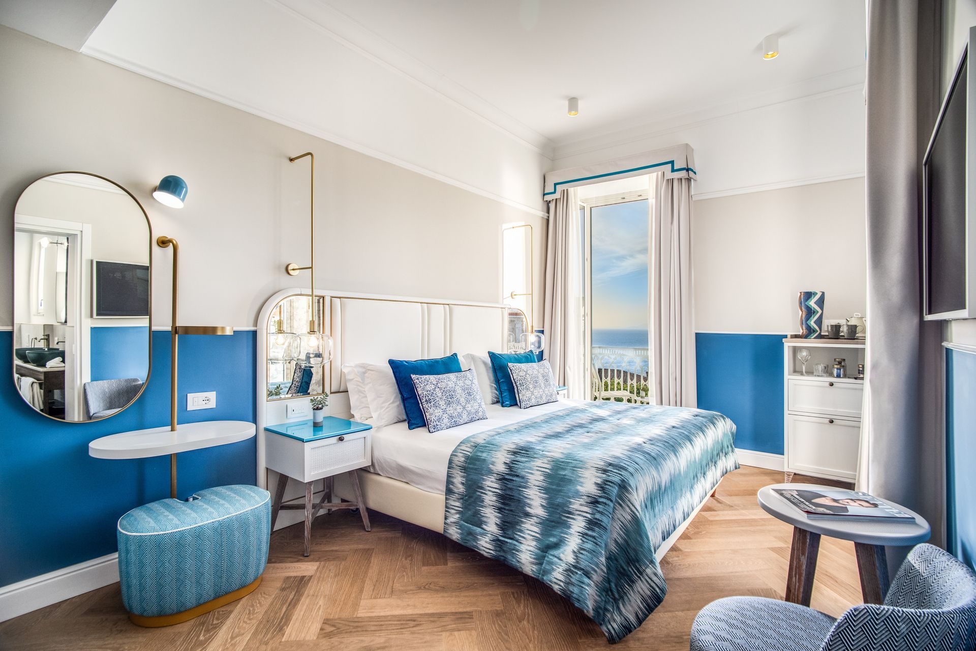 Una luminosa camera d'albergo con letto matrimoniale, lenzuola con motivi blu, pareti blu, specchio da trucco e balcone con vista mare.