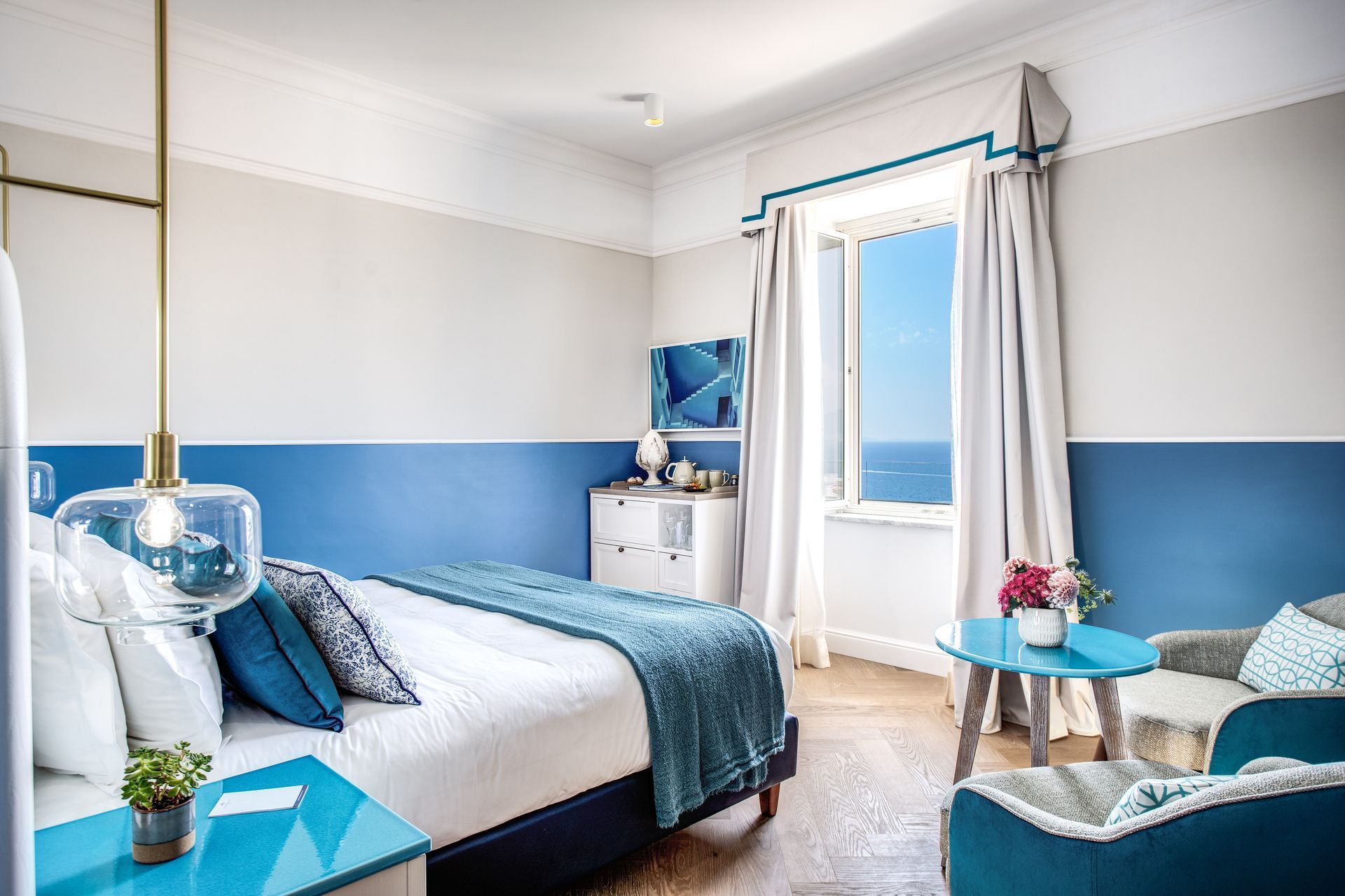 Una camera d'albergo con un letto bianco, dettagli blu e una finestra con vista sull'oceano.