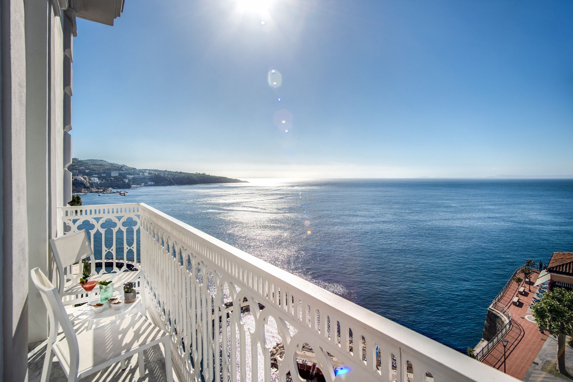 White balcony with a small table and chairs overlooking the blue Mediterranean sea under a bright, sunny sky.