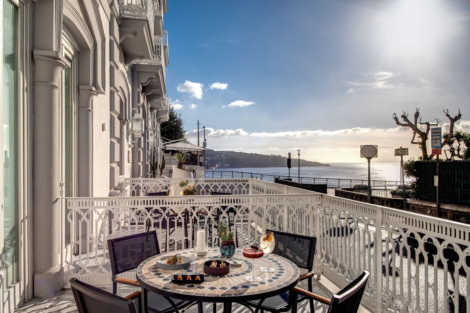 Un tavolino da bistrot per due persone su un balcone bianco e decorato, affacciato su una baia soleggiata.