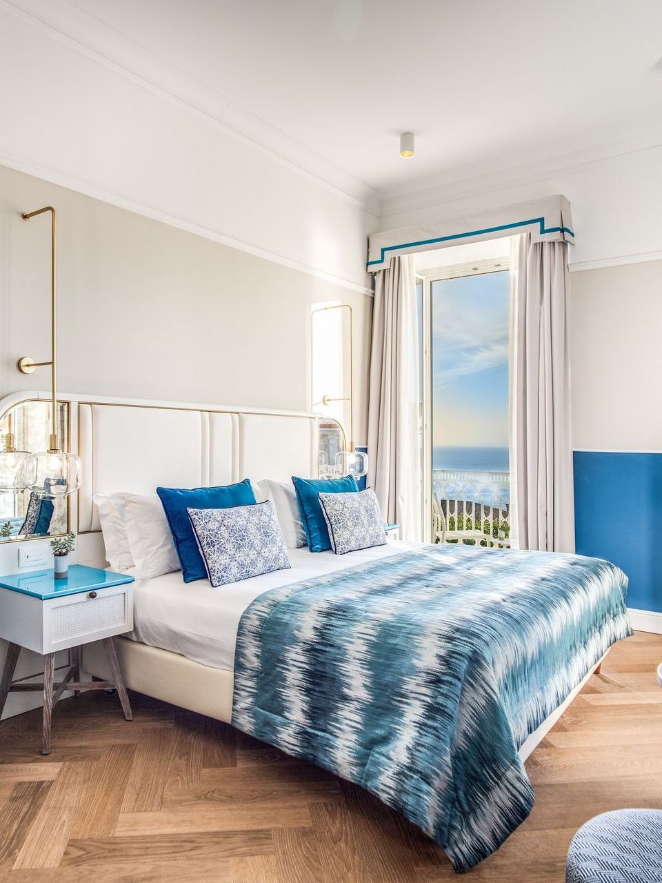 A sunlit bedroom with a blue and white patterned bedspread, matching pillows, and a window overlooking the ocean.