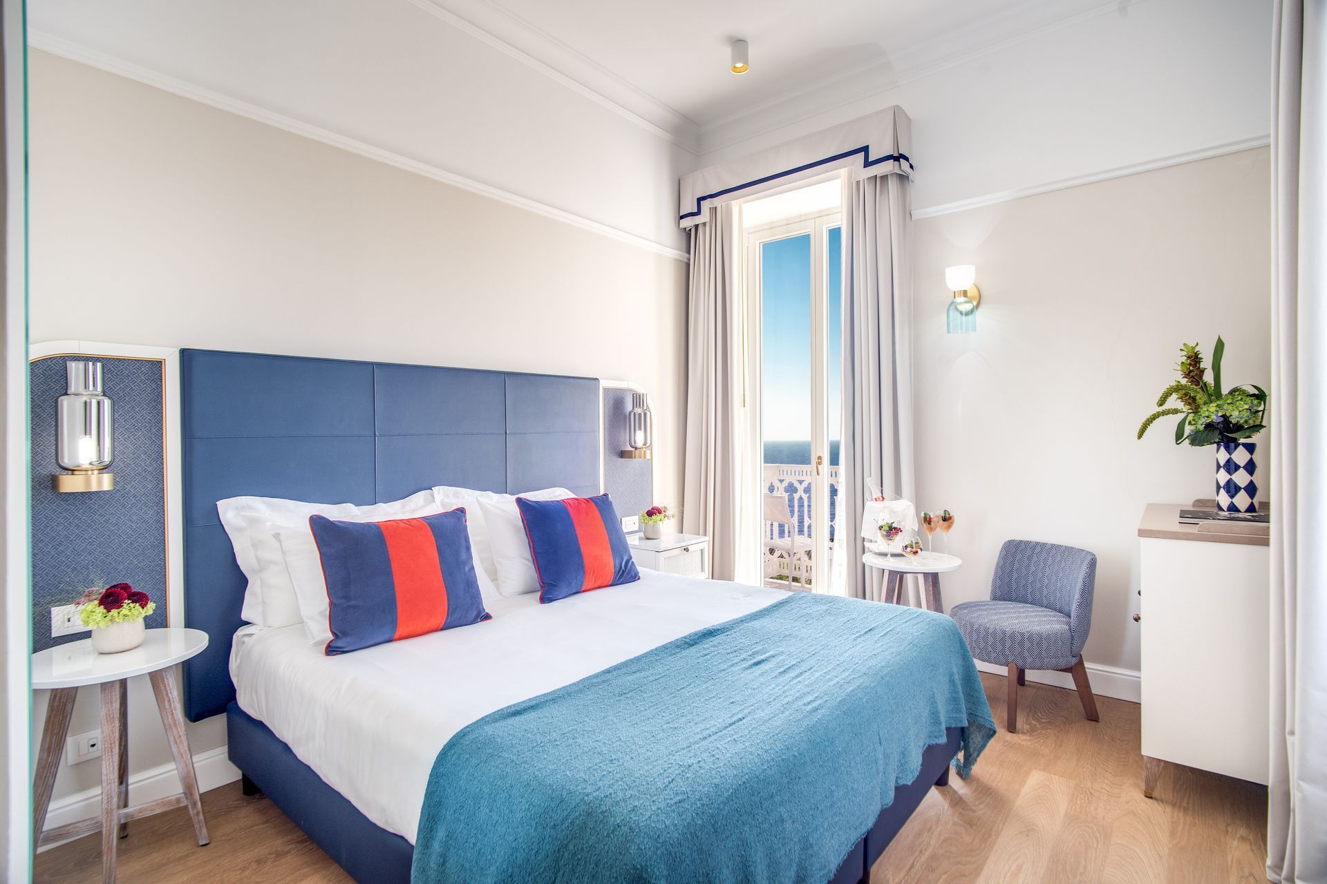 A hotel room with a blue upholstered bed, matching throw blanket, white bedding, a small chair, and a balcony view.