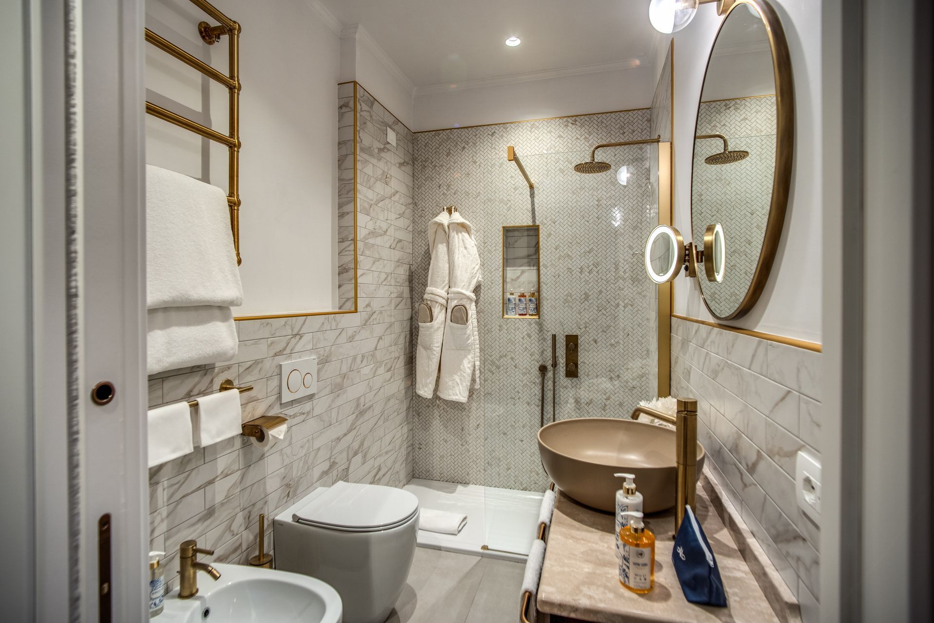 Bagno moderno con pareti in marmo, lavabo a colonna, WC, cabina doccia con accappatoi e specchio con cornice dorata.