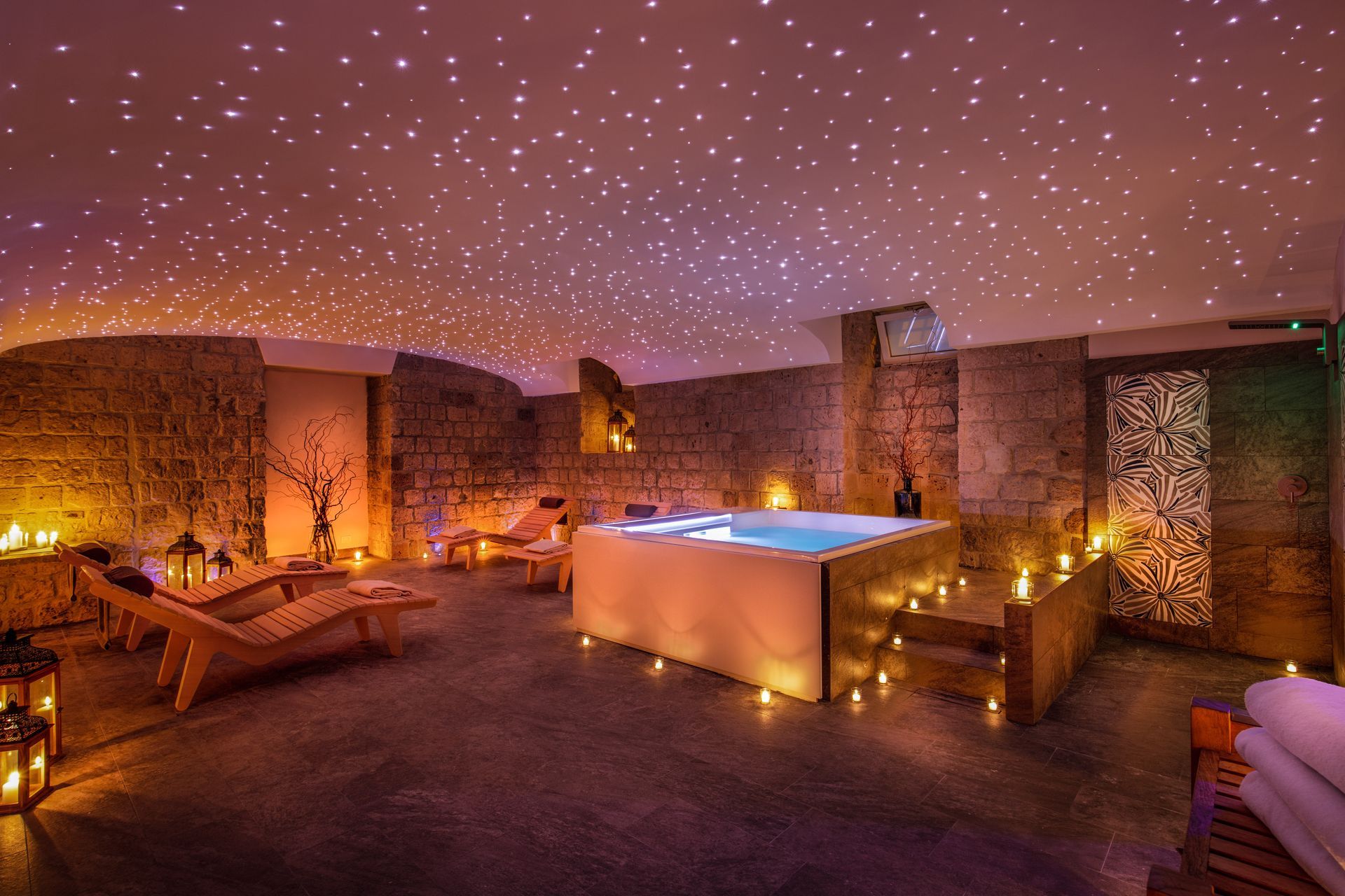 A luxurious spa room with stone walls, a central hot tub, lounge chairs, and a star-lit ceiling with warm candle lighting.